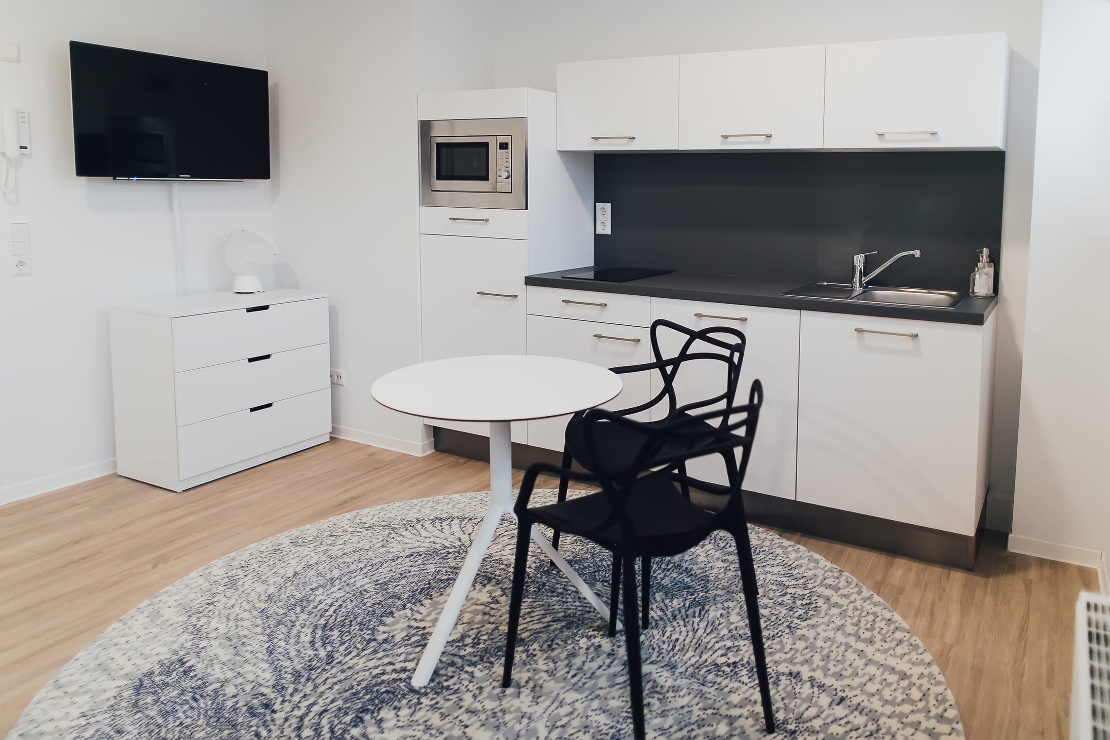 Wohn-/Küchenbereich mit heller Einbauküche, runder Tisch mit zwei Stühlen, ovaler Teppich
