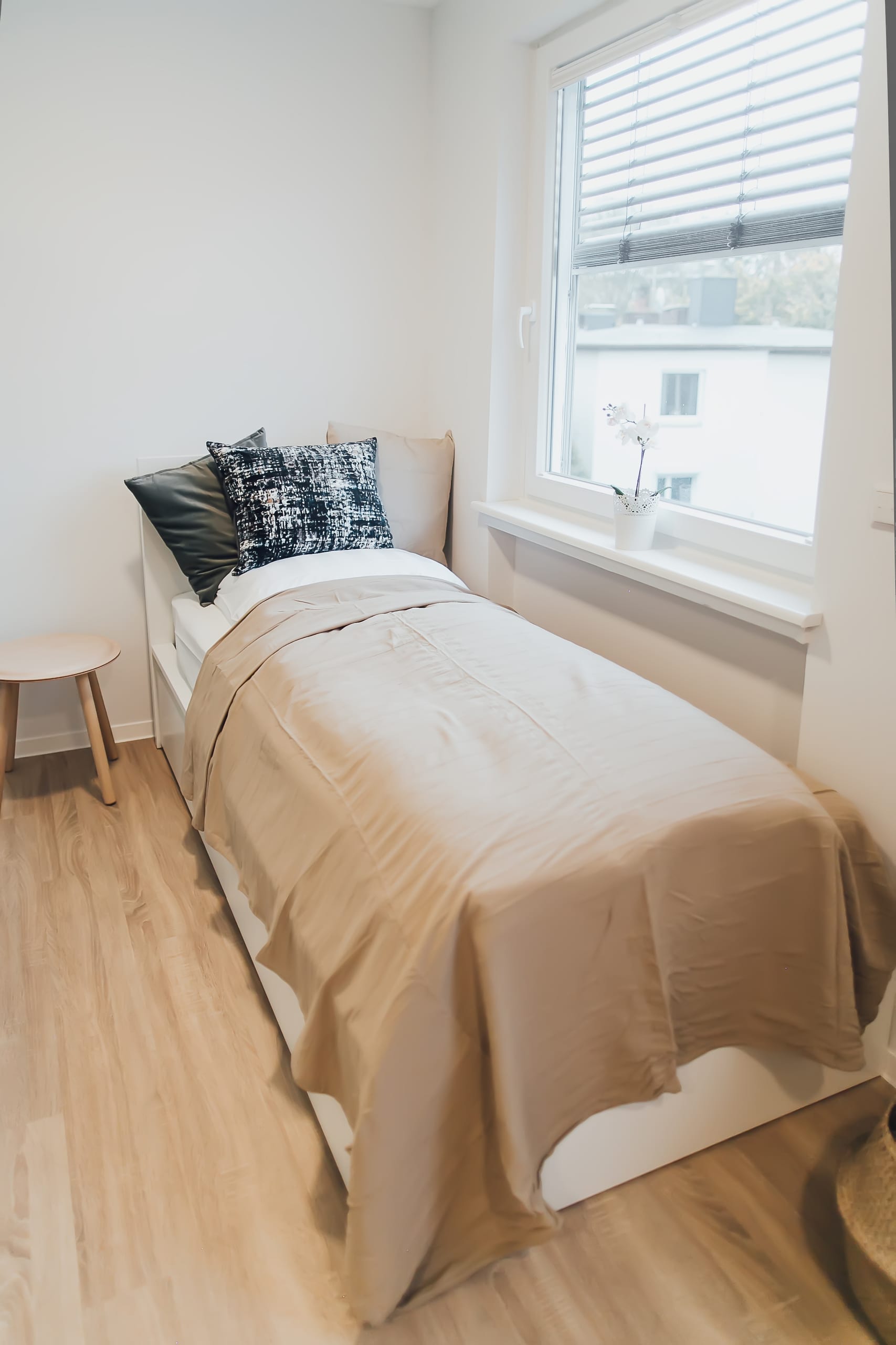 Schlafzimmer mit weißem Bettrahmen, beigefarbener Decke, drei Kissen, Holzhocker und Fenster