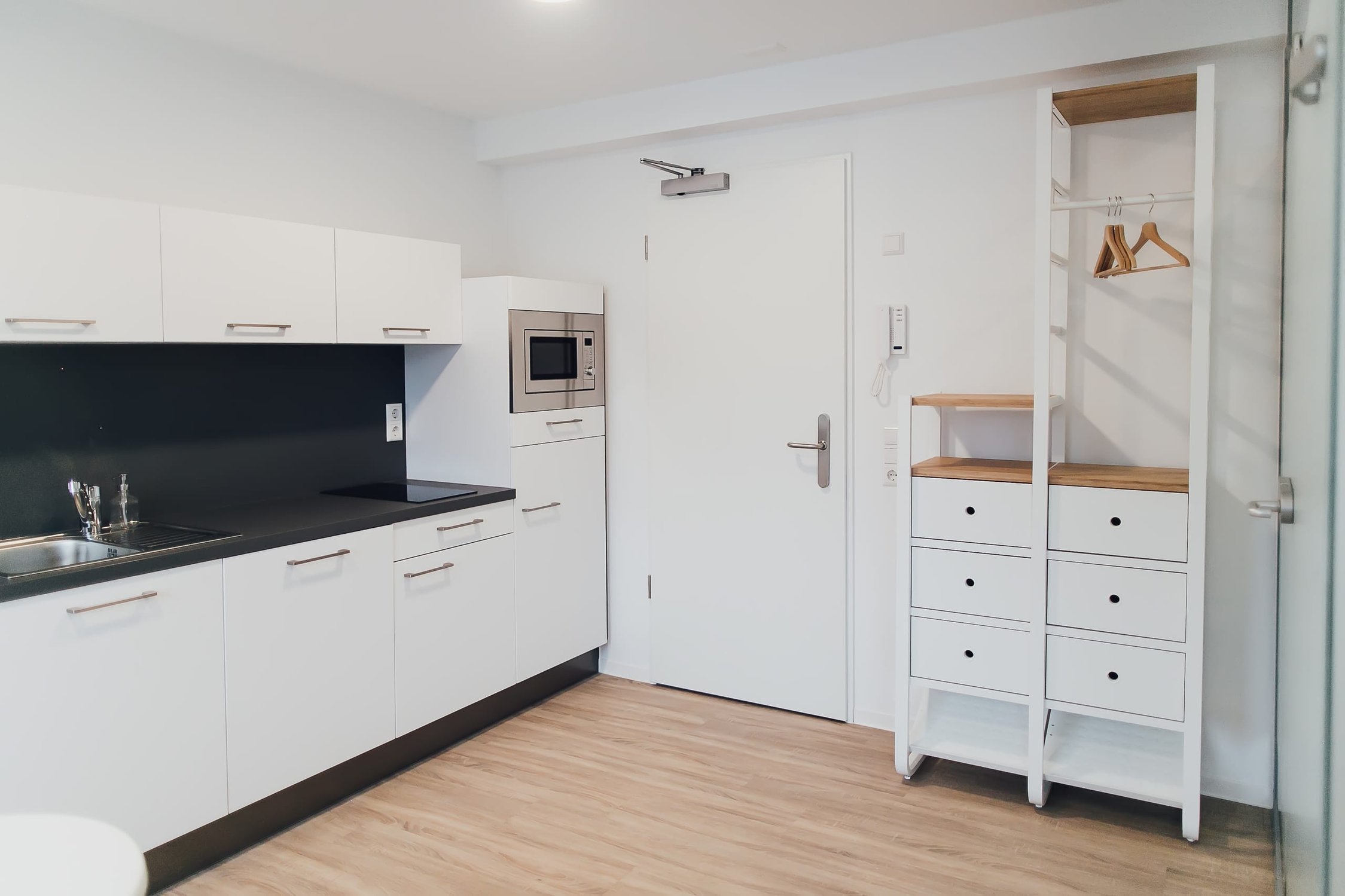 Helle Küchenzeile mit weißer Front, schwarzer Arbeitsplatte und integrierter Mikrowelle, daneben offener Schrank