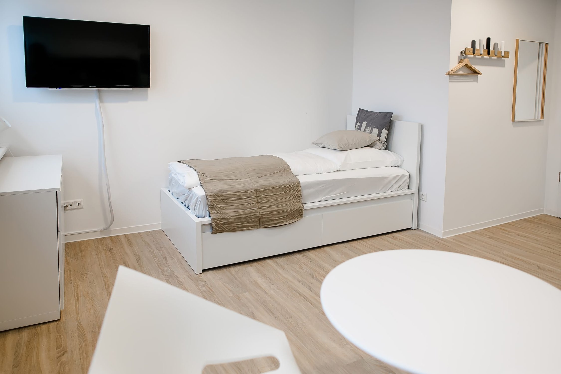 Heller Raum mit Einzelbett, weißem Nachttisch, TV an Wand, Holzboden