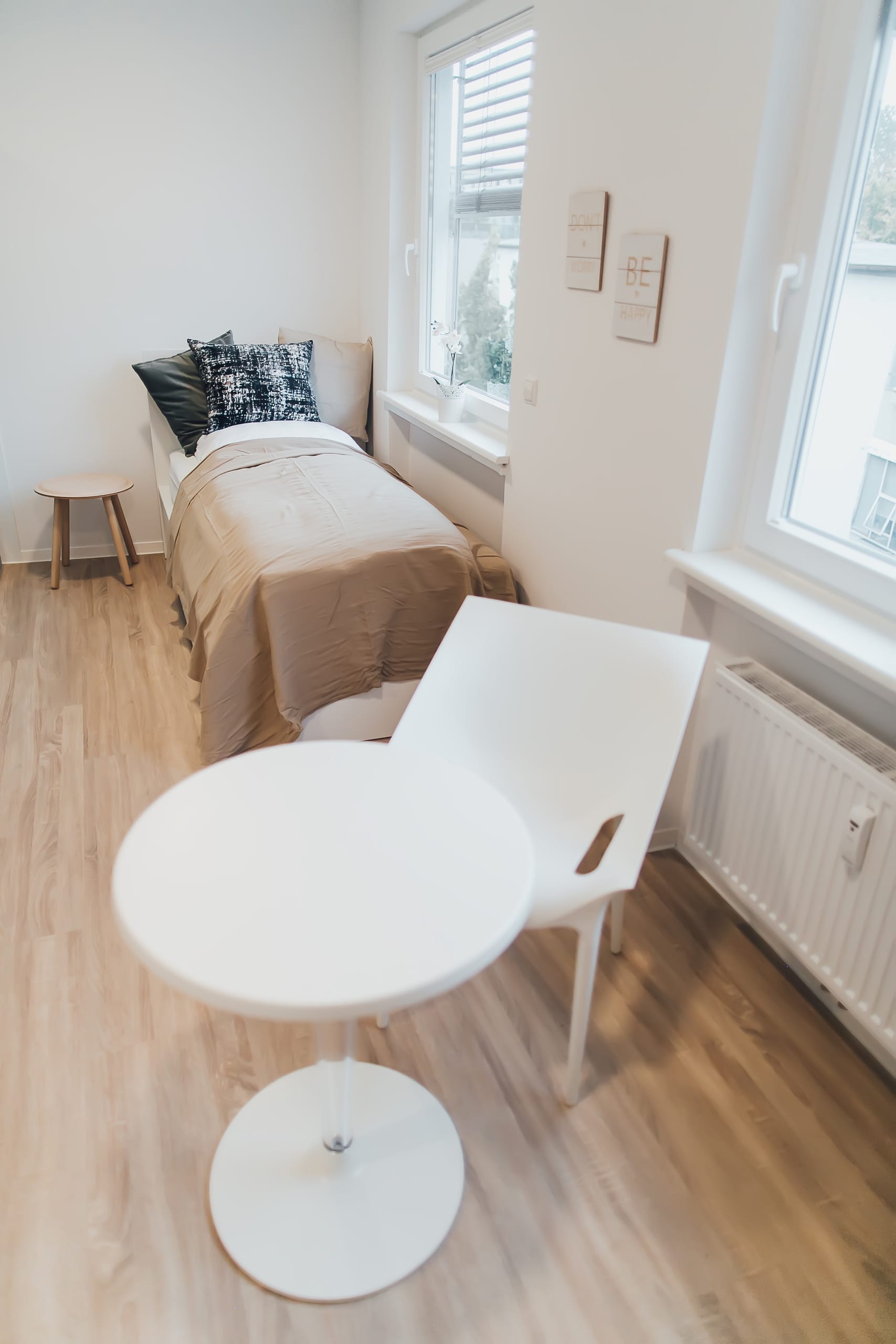 Helles Zimmer mit Bett, kleinem Holztisch, großem Fenster und weißem Bistrotisch mit Stuhl