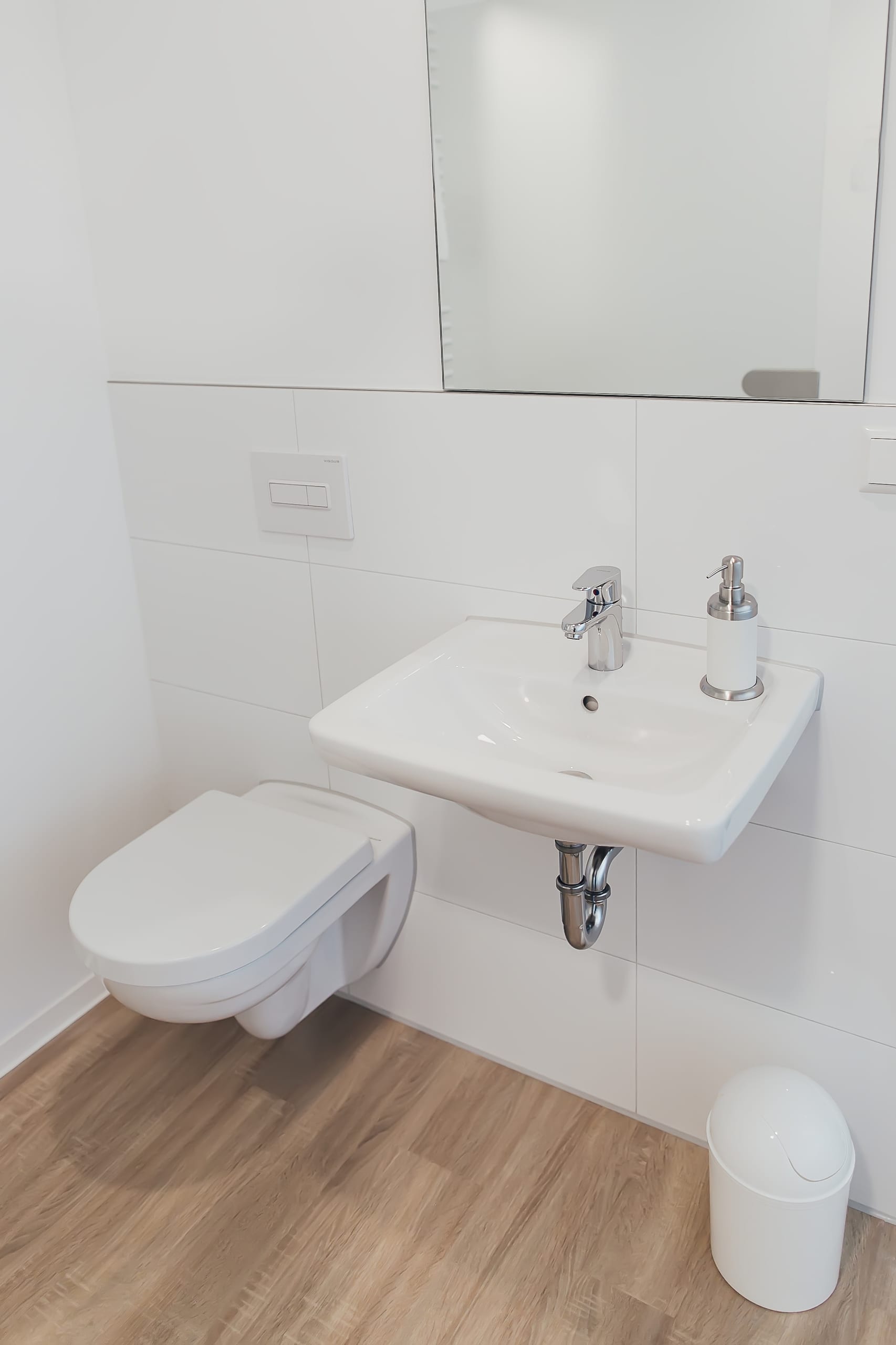 Modernes Bad mit weißem Waschbecken und Toilette, Wandfliesen, heller Holzboden, weißer Mülleimer