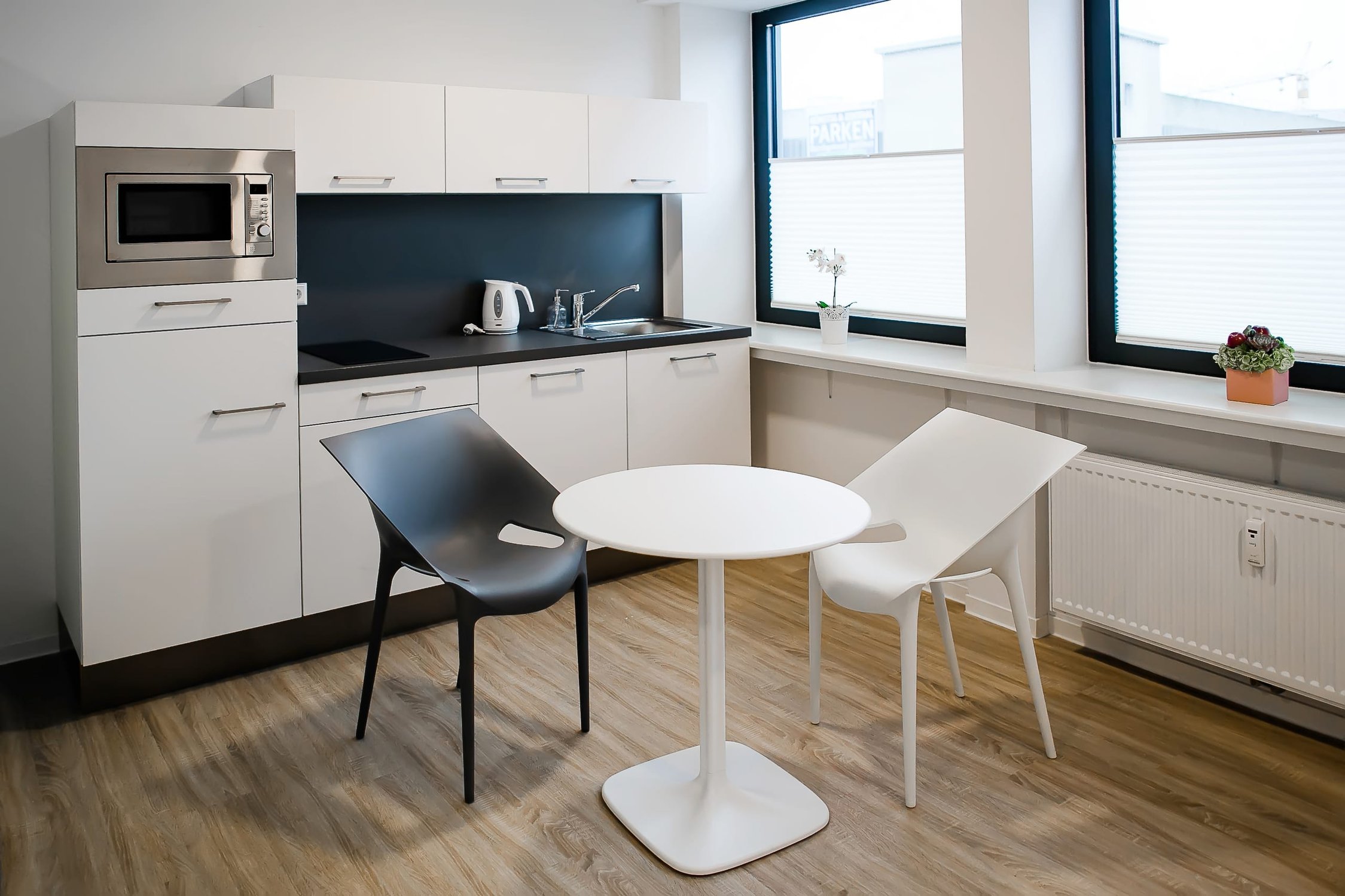 Küchenzeile mit weißen Schränken, Mikrowelle, Spüle, hellem Holzboden und zwei Stühlen am Tisch