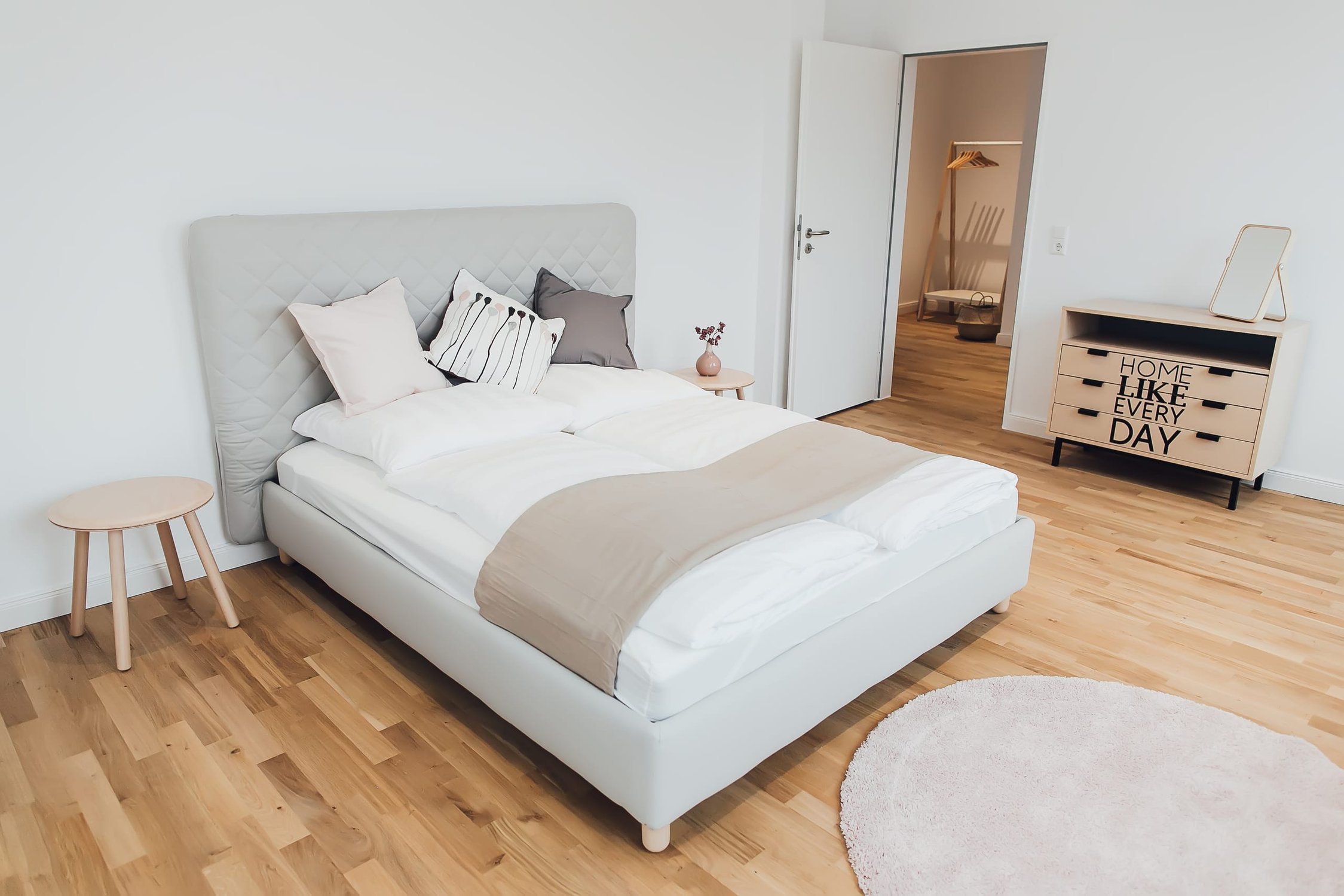 Helles Schlafzimmer mit grauem Bett, Holzboden und Kommode, Tür zu weiterem Raum