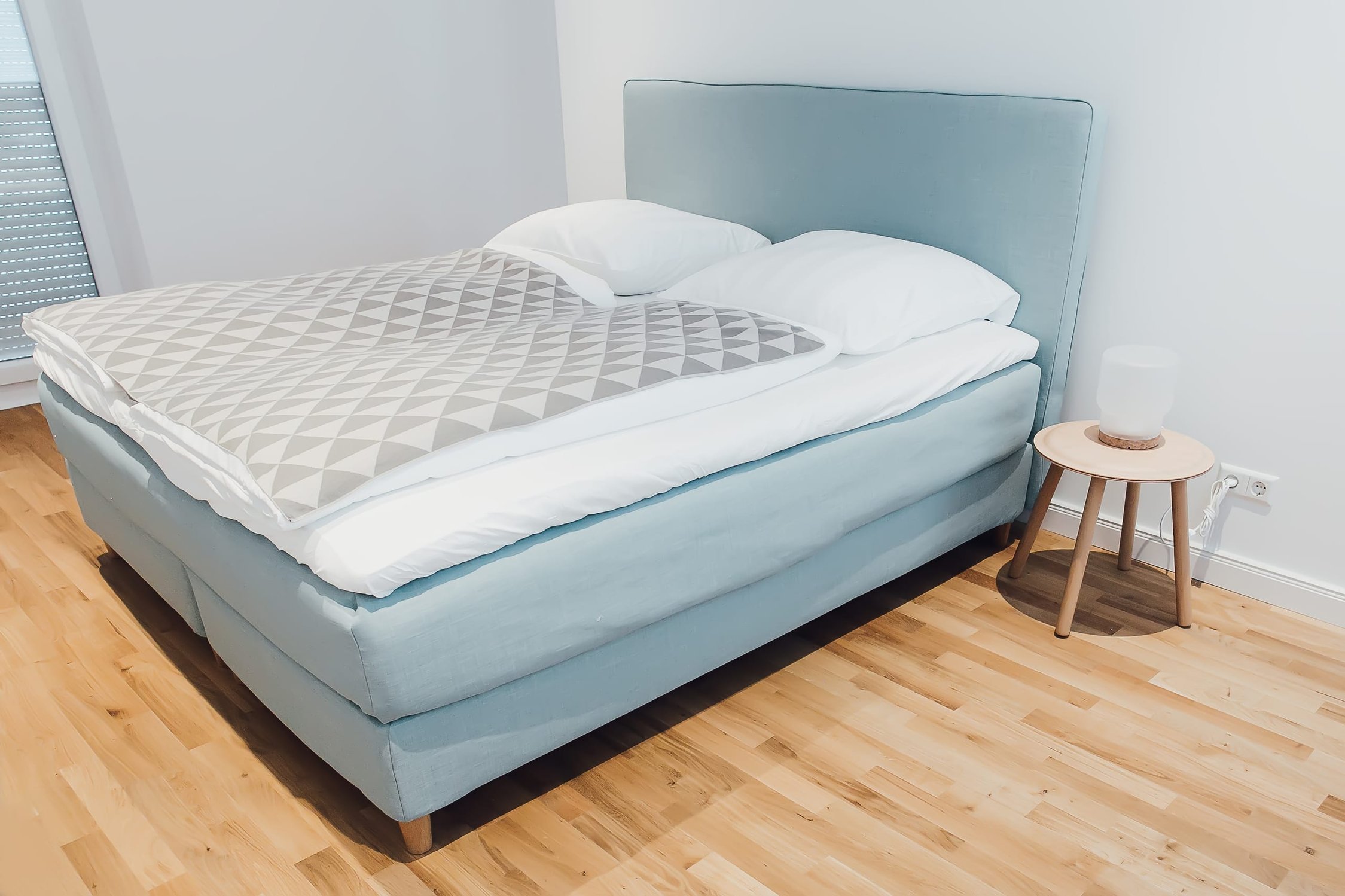 Schlafzimmer mit hellblauem Boxspringbett, hellem Holzboden und kleinem Nachttisch