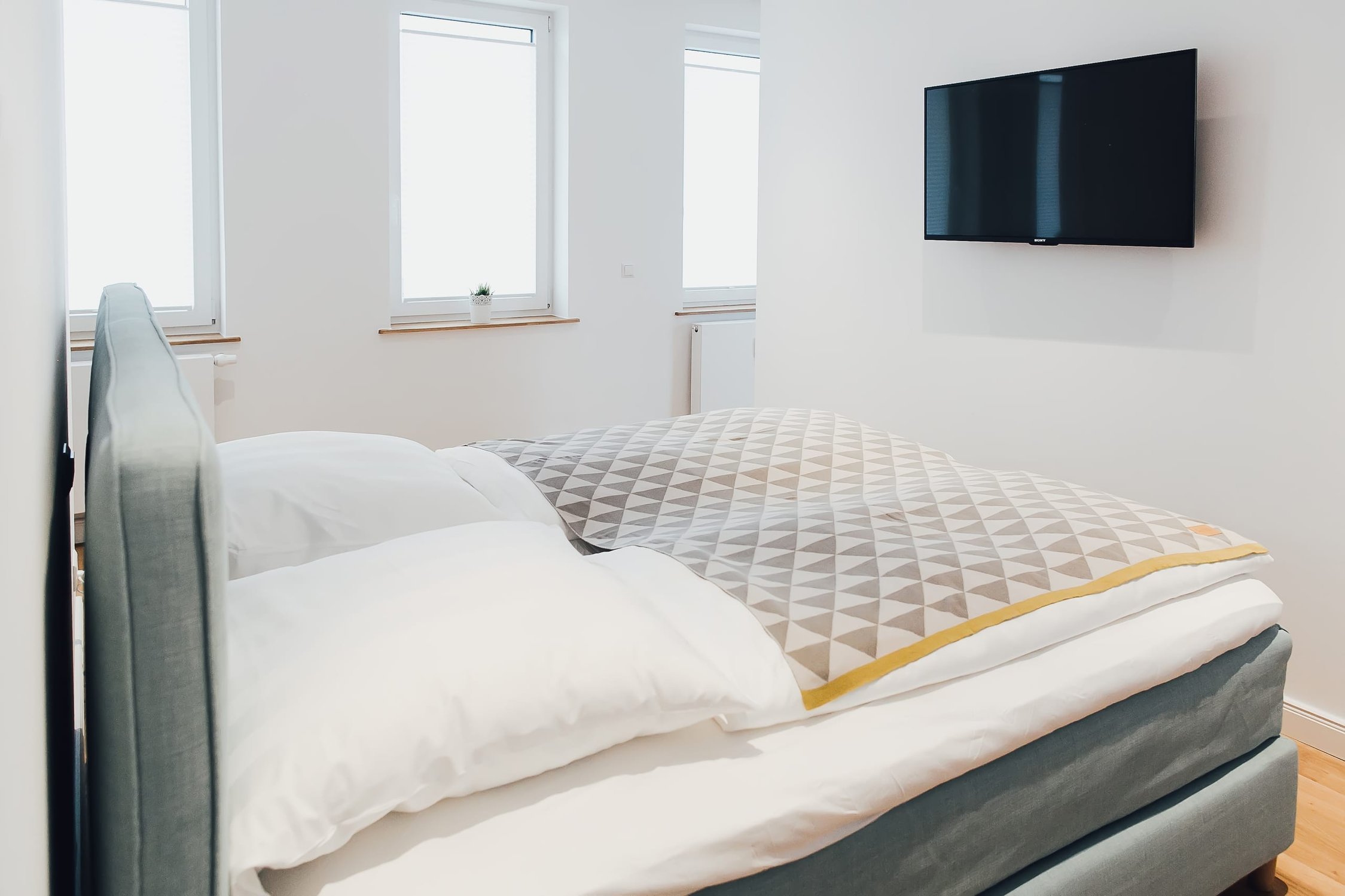 Schlafzimmer mit Doppelbett, zwei Fenstern und Fernseher an weißer Wand