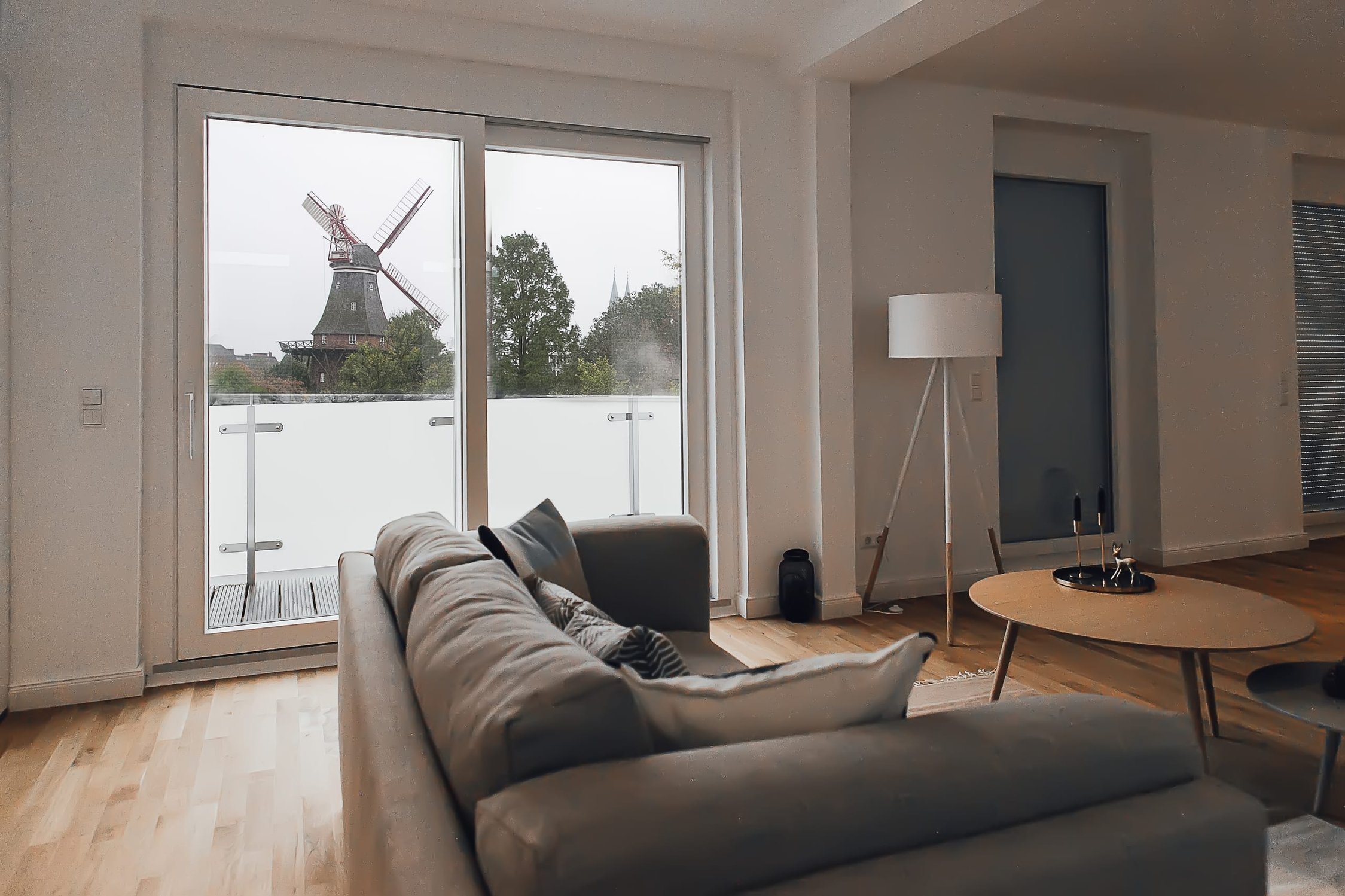 Wohnzimmer mit grauem Sofa, hellem Holzboden und Blick auf Windmühle und Baumlandschaft