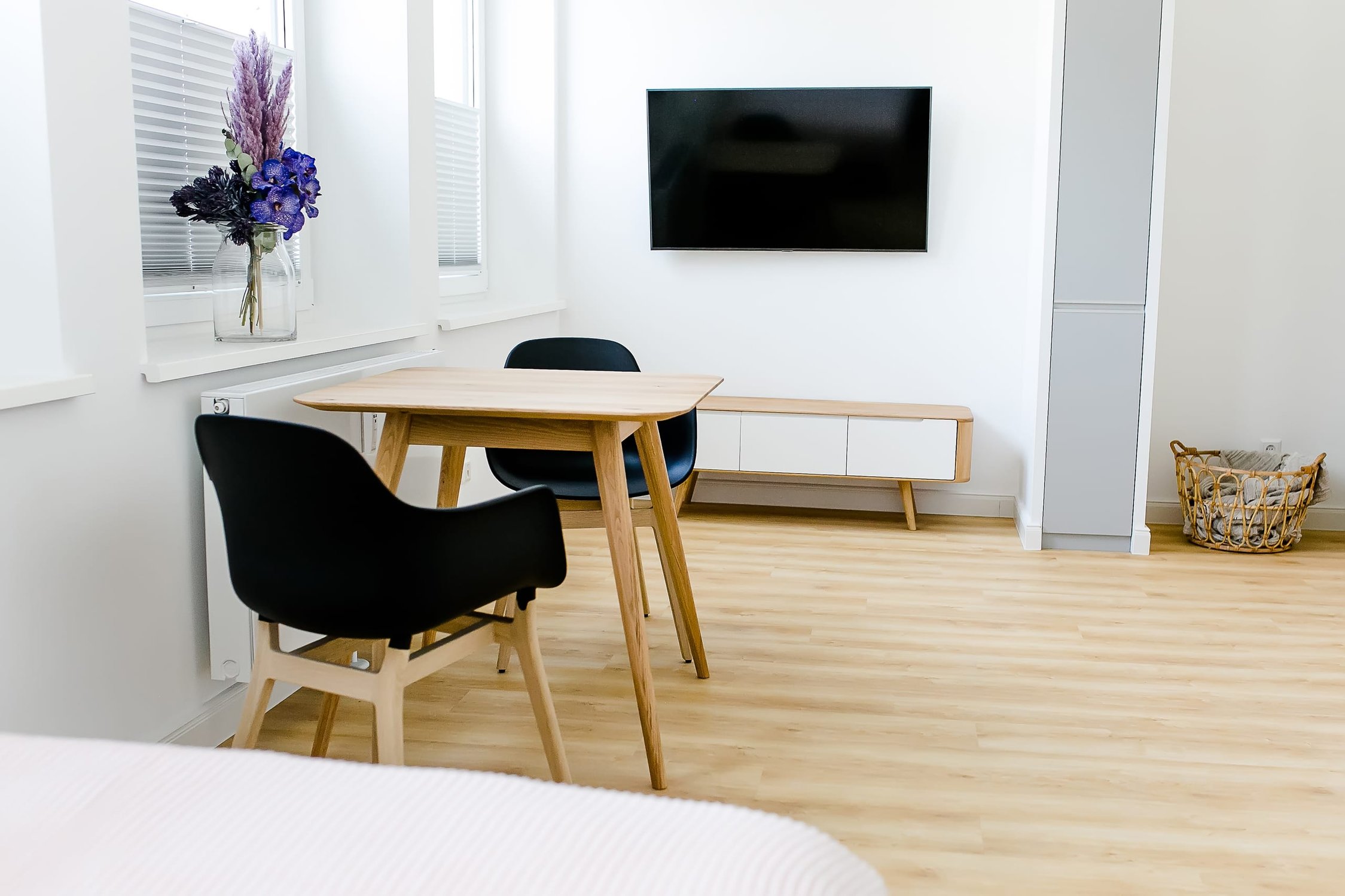 Helles Zimmer mit Holzboden, Esstisch mit schwarzen Stühlen, TV und Blumenvase auf Fensterbank