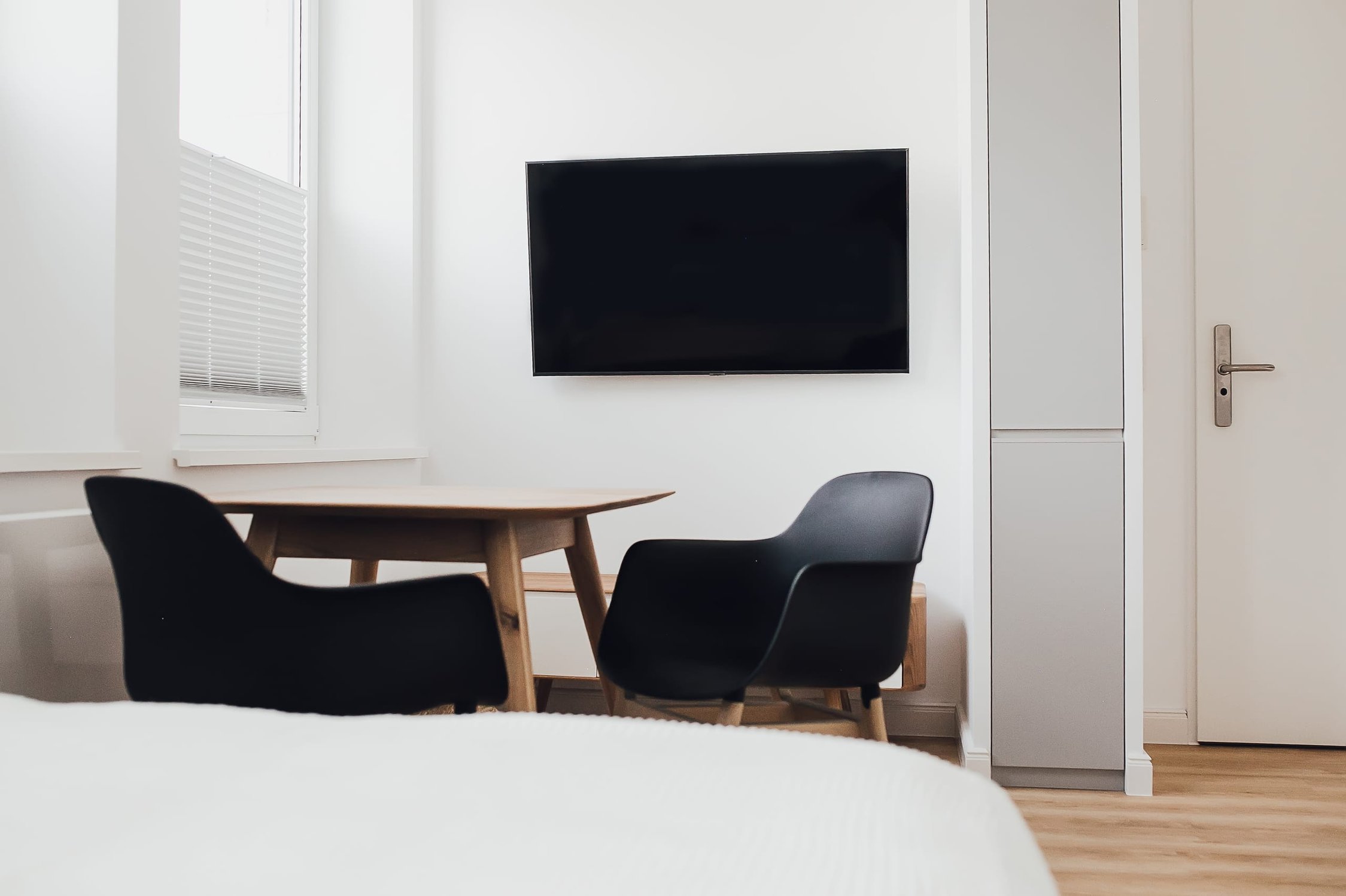 Helles Zimmer mit kleinem Holztisch, schwarzen Stühlen, weißer Wand und Flachbildfernseher