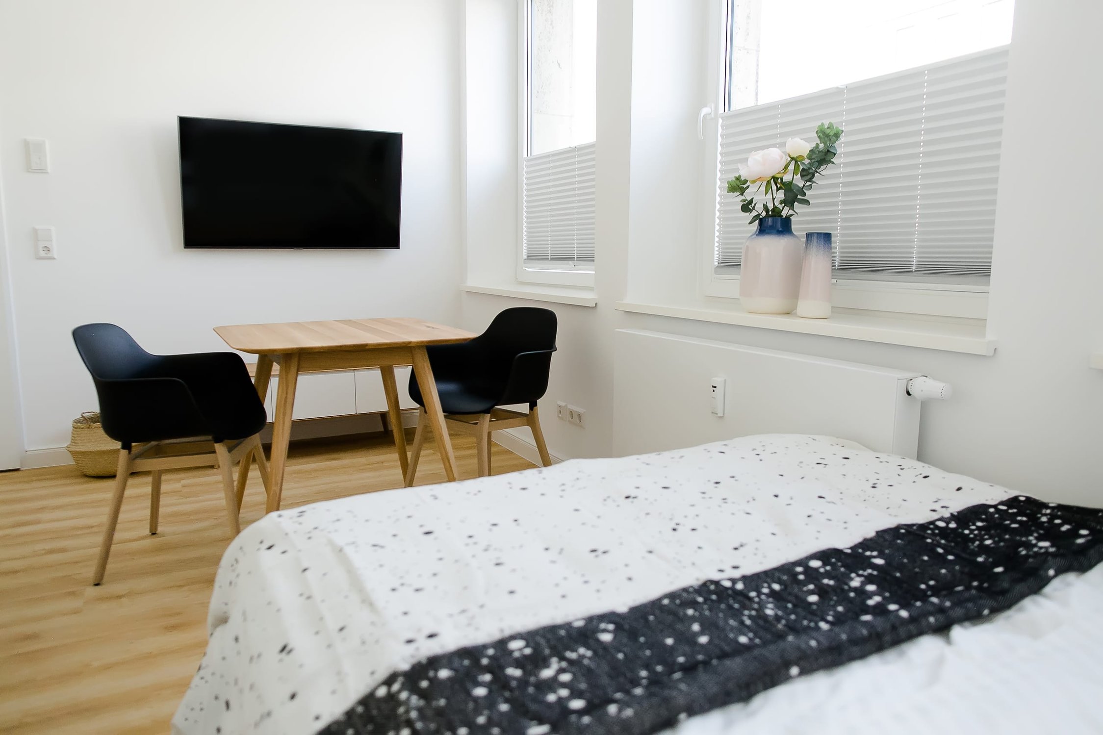 Helles Schlafzimmer mit Smart-TV, Holztisch, schwarzen Stühlen, weißer Bettwäsche, Fensterbank-Deko