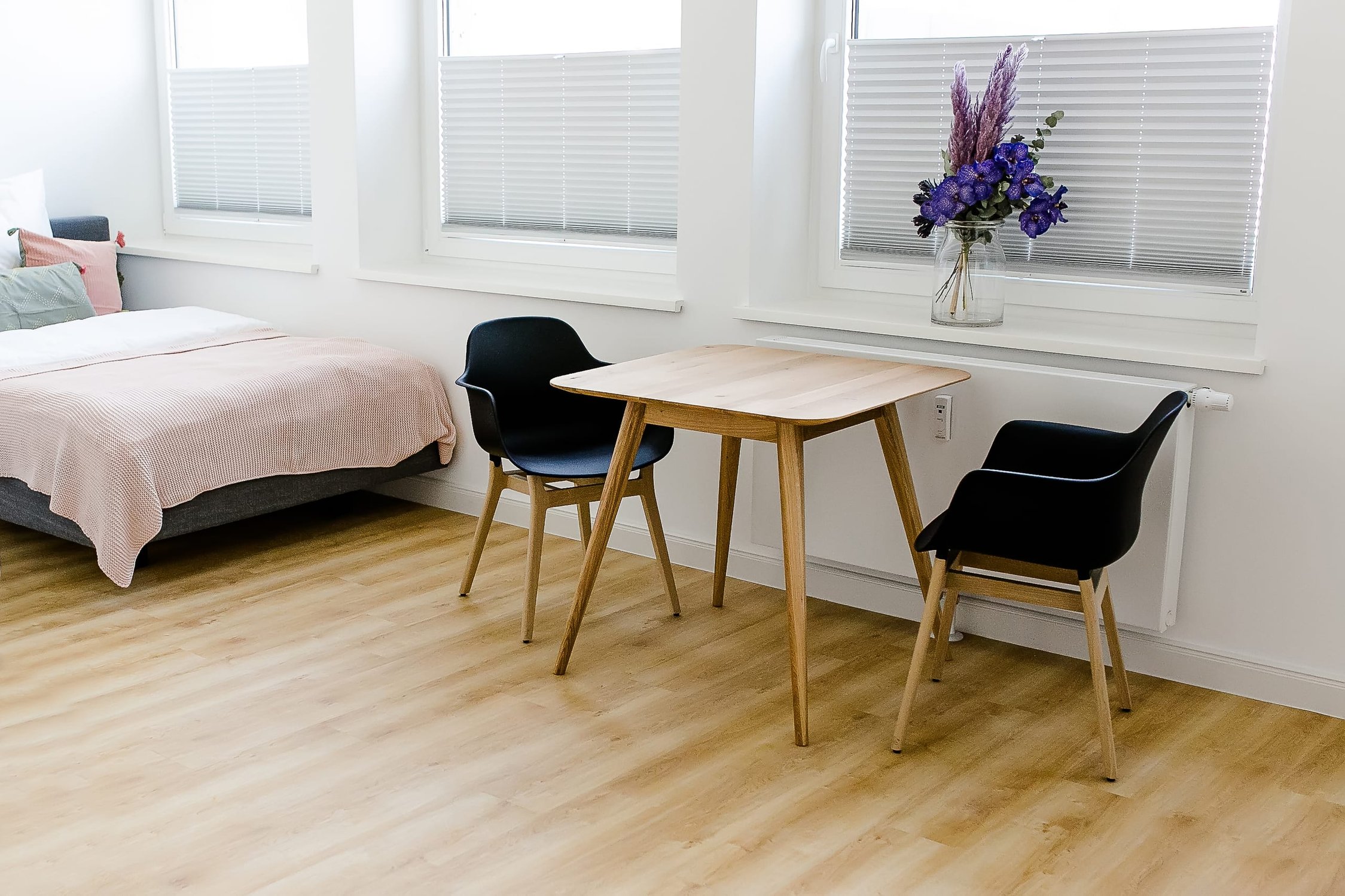 Heller Raum mit Bett, Holztisch, zwei schwarzen Stühlen, zwei Fenstern, Blumenstrauß