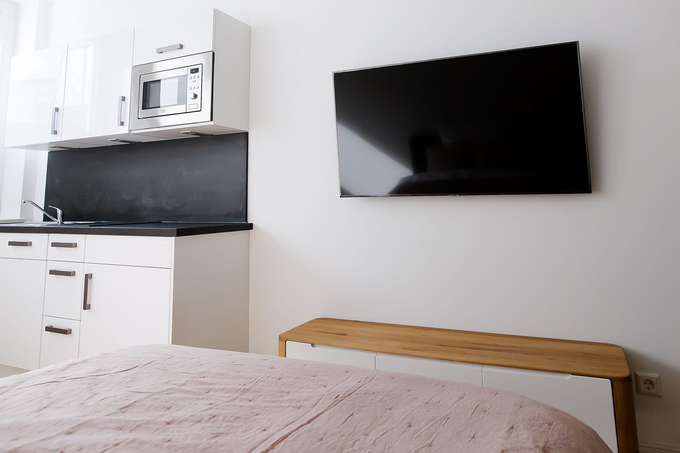 Kleine Wohnküche mit weißen Schränken, Mikrowelle, Spüle und schwarzer Arbeitsplatte, daneben TV über Holzbank