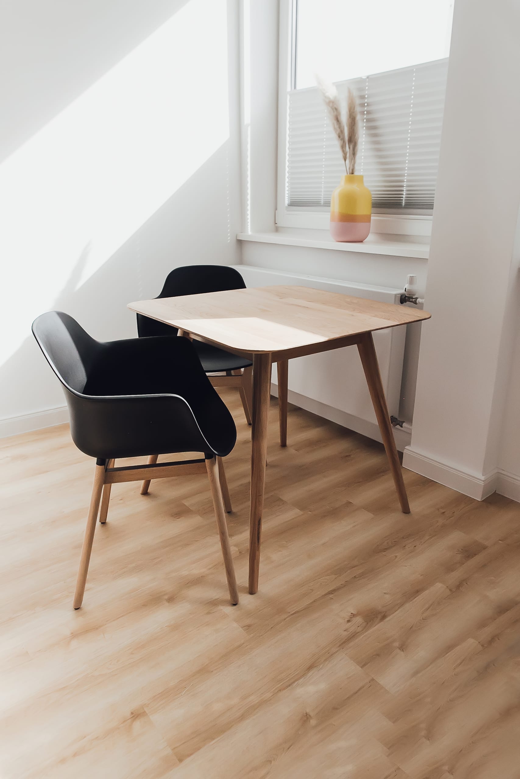 Kleiner Esstisch aus hellem Holz mit zwei schwarzen Stühlen, vor Fenster mit Jalousie