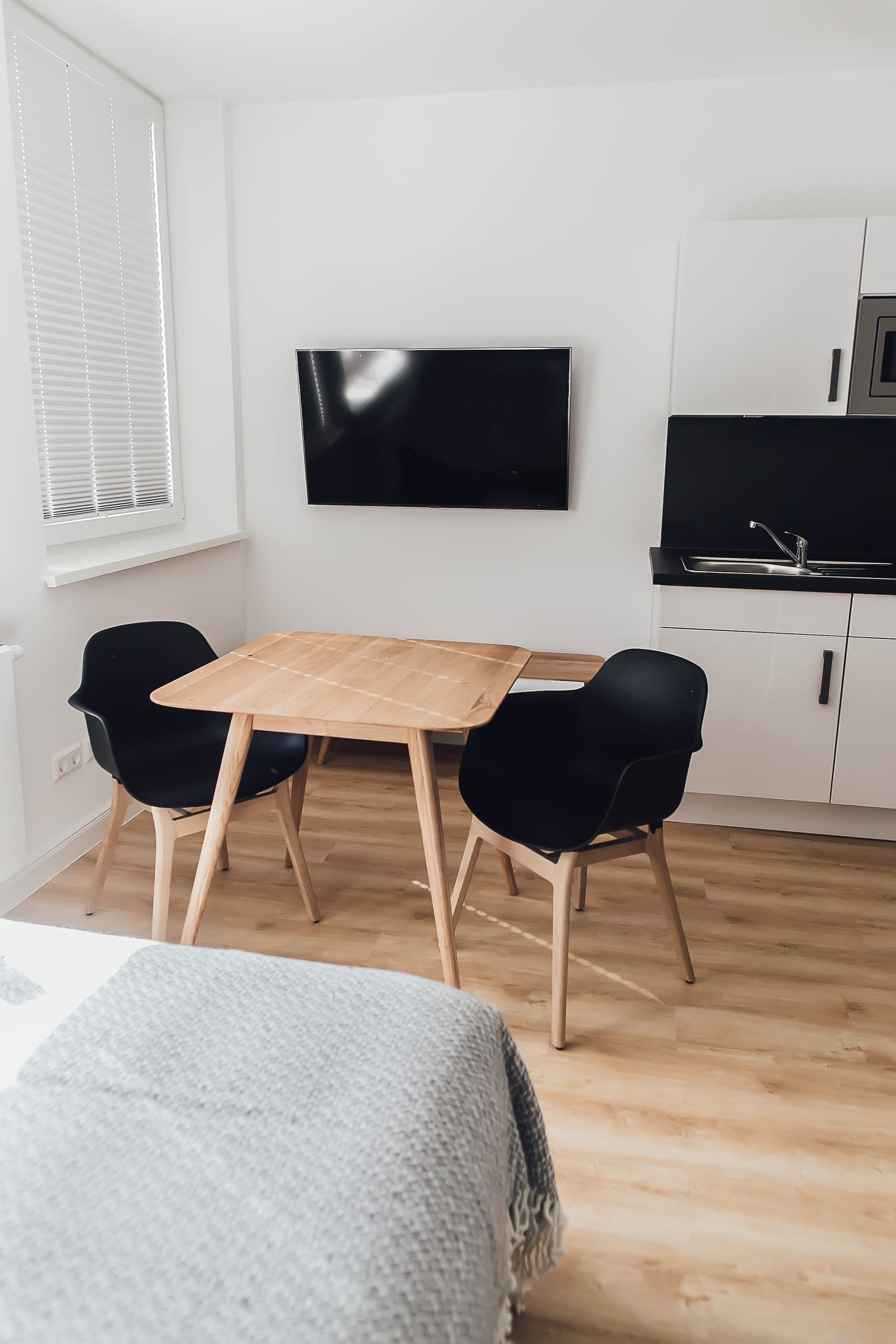 Kleiner Wohnraum mit Holztisch, schwarzen Stühlen, Fernseher und weißer Küchenzeile