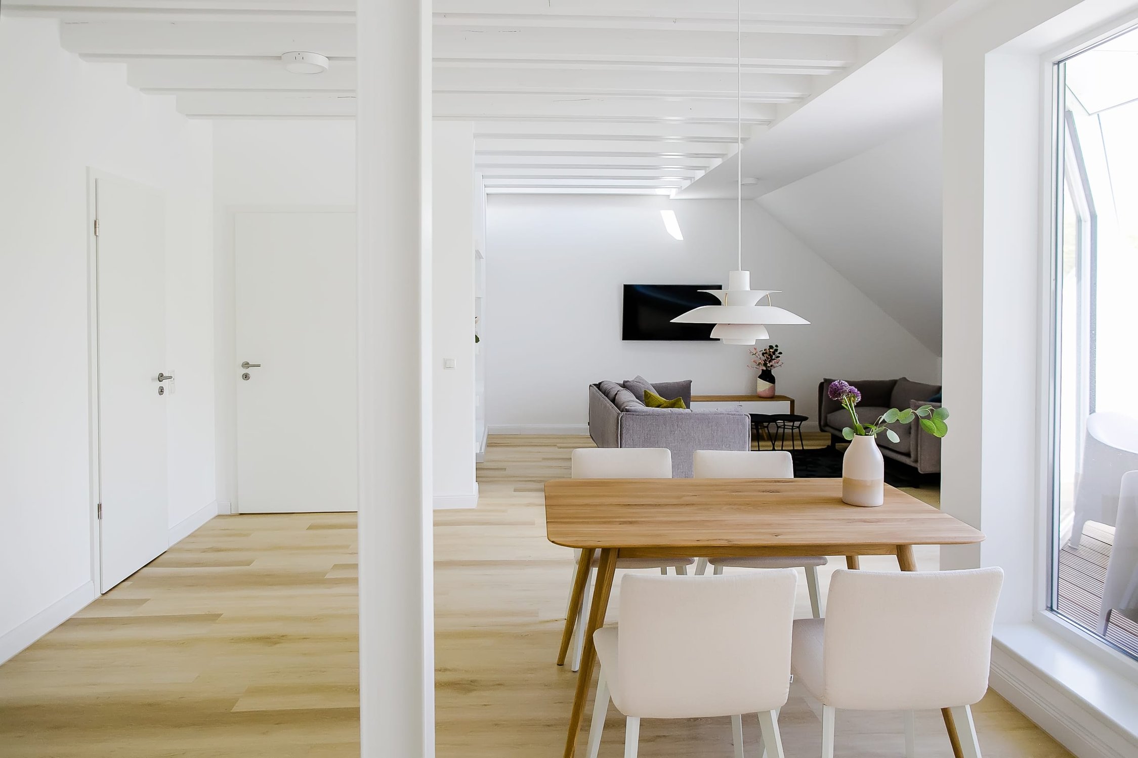 Helles, offenes Wohnzimmer mit Holzboden, Esstisch und Stühlen, im Hintergrund ein Sofa und Fernseher