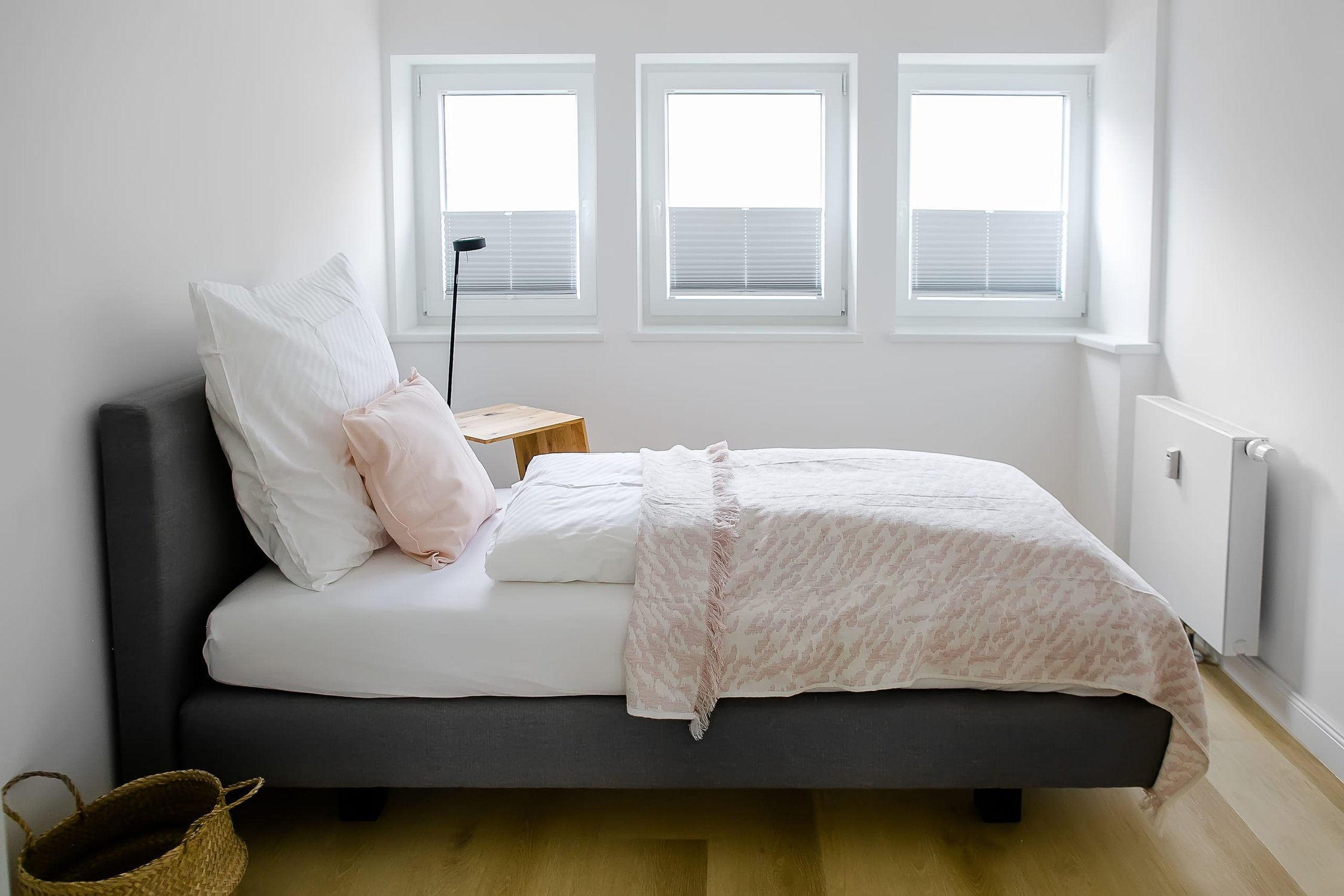 Schlafzimmer mit grauem Bett, weißen Kissen und drei Fenstern mit Jalousien, heller Holzboden