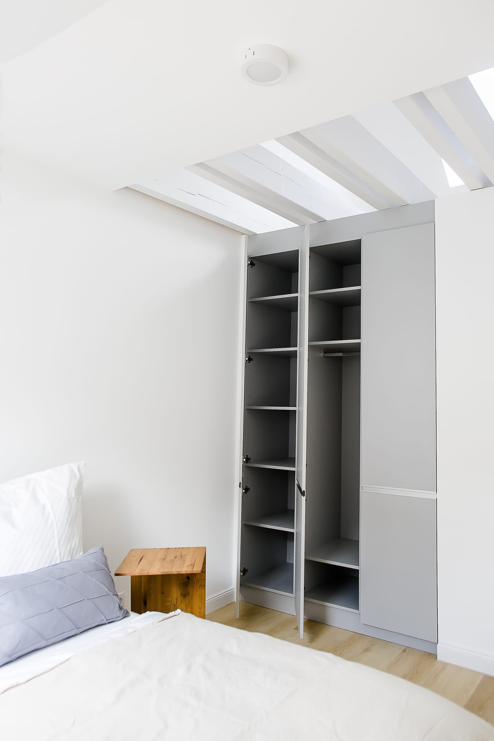 Schlafzimmer mit weißer Bettwäsche, hölzernem Nachttisch und offenem grauen Kleiderschrank