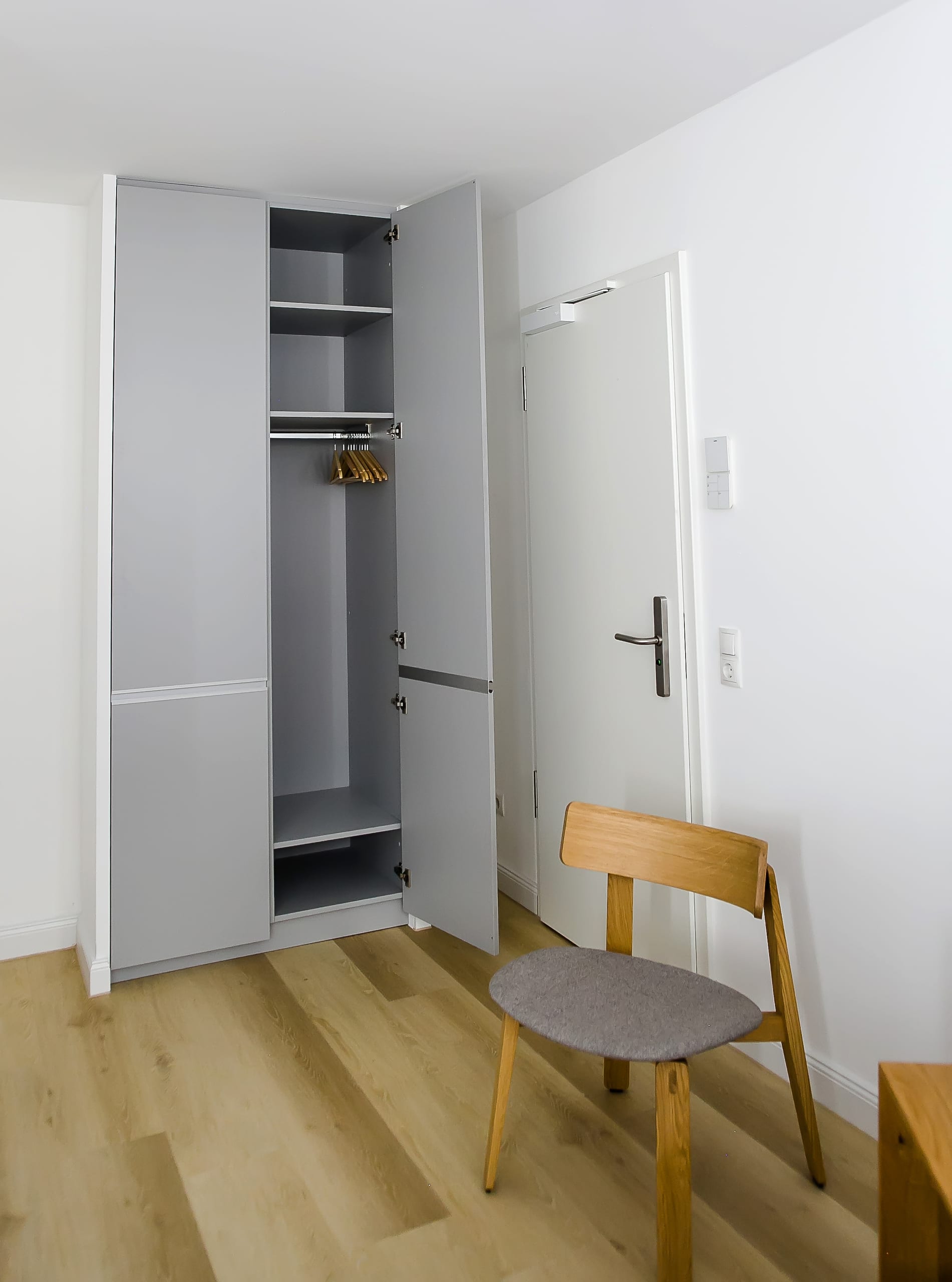 Grauer offener Schrank mit Kleiderbügeln und hellem Holzboden, flankiert von einem Stuhl