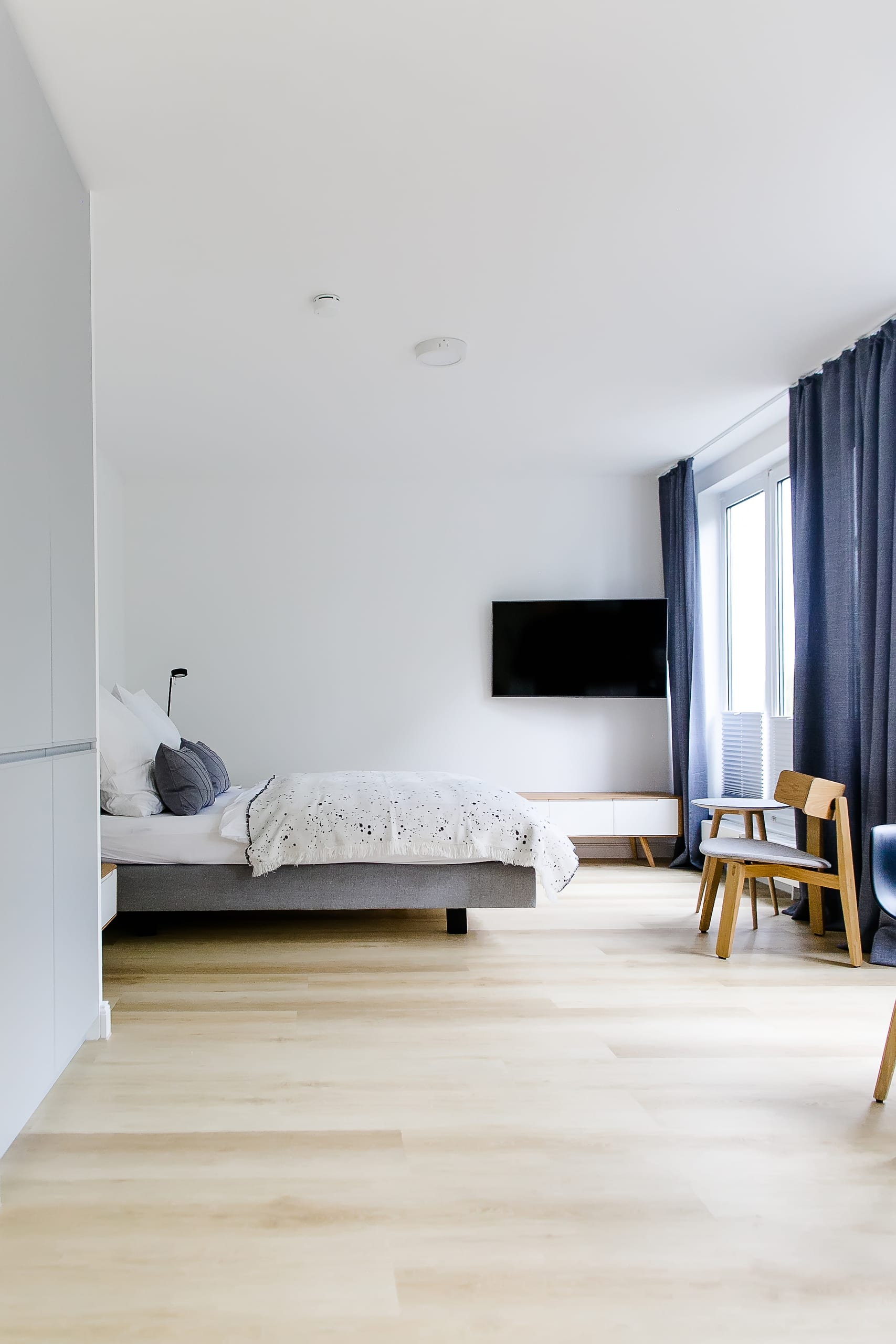 Modernes Schlafzimmer mit hellem Holzboden, grauem Bett und großem Fenster mit blauen Vorhängen