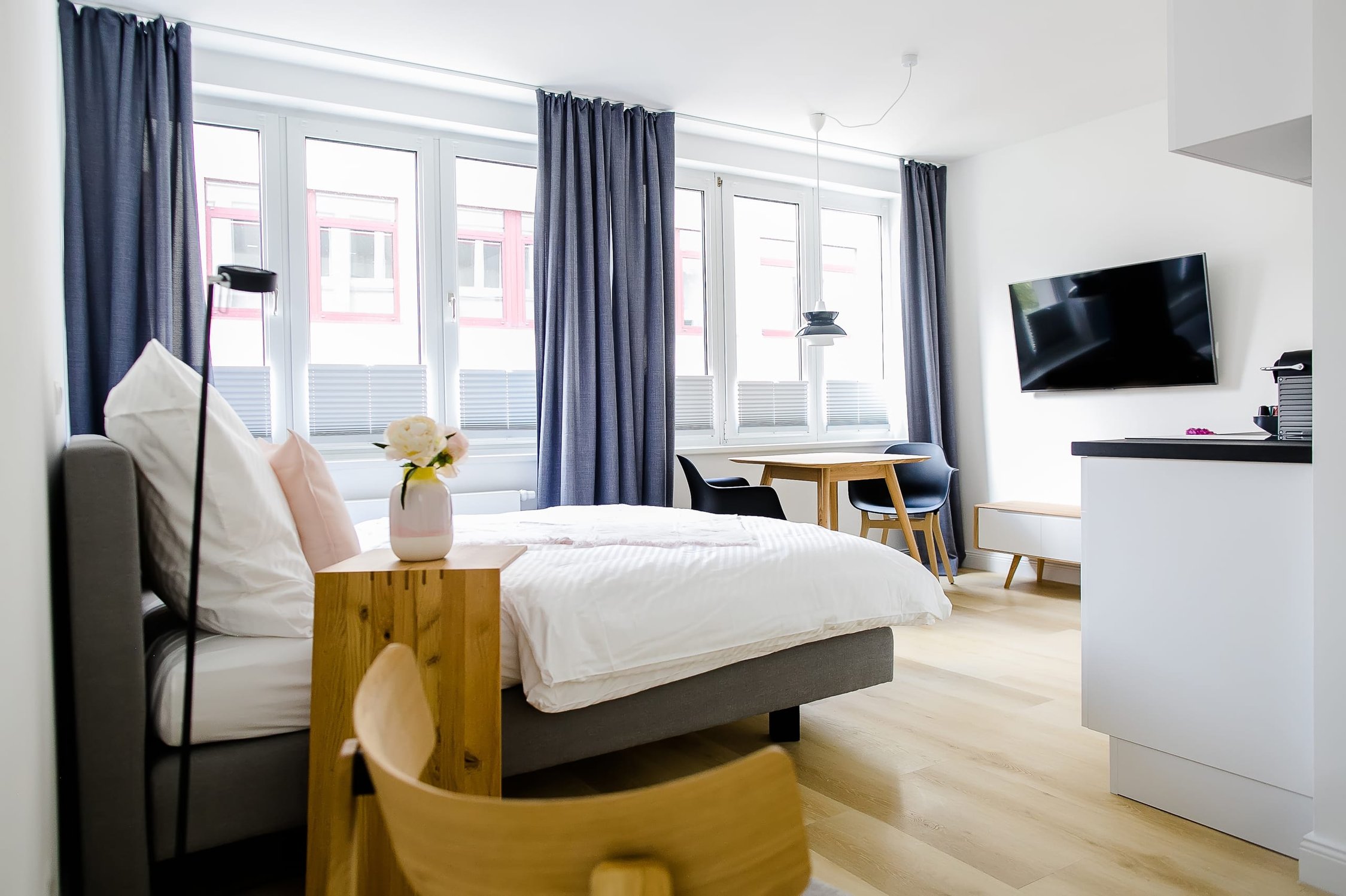 Helles Apartment mit großem Bett, Fernseher und Essbereich am Fenster, Holz- und Grau-Akzente