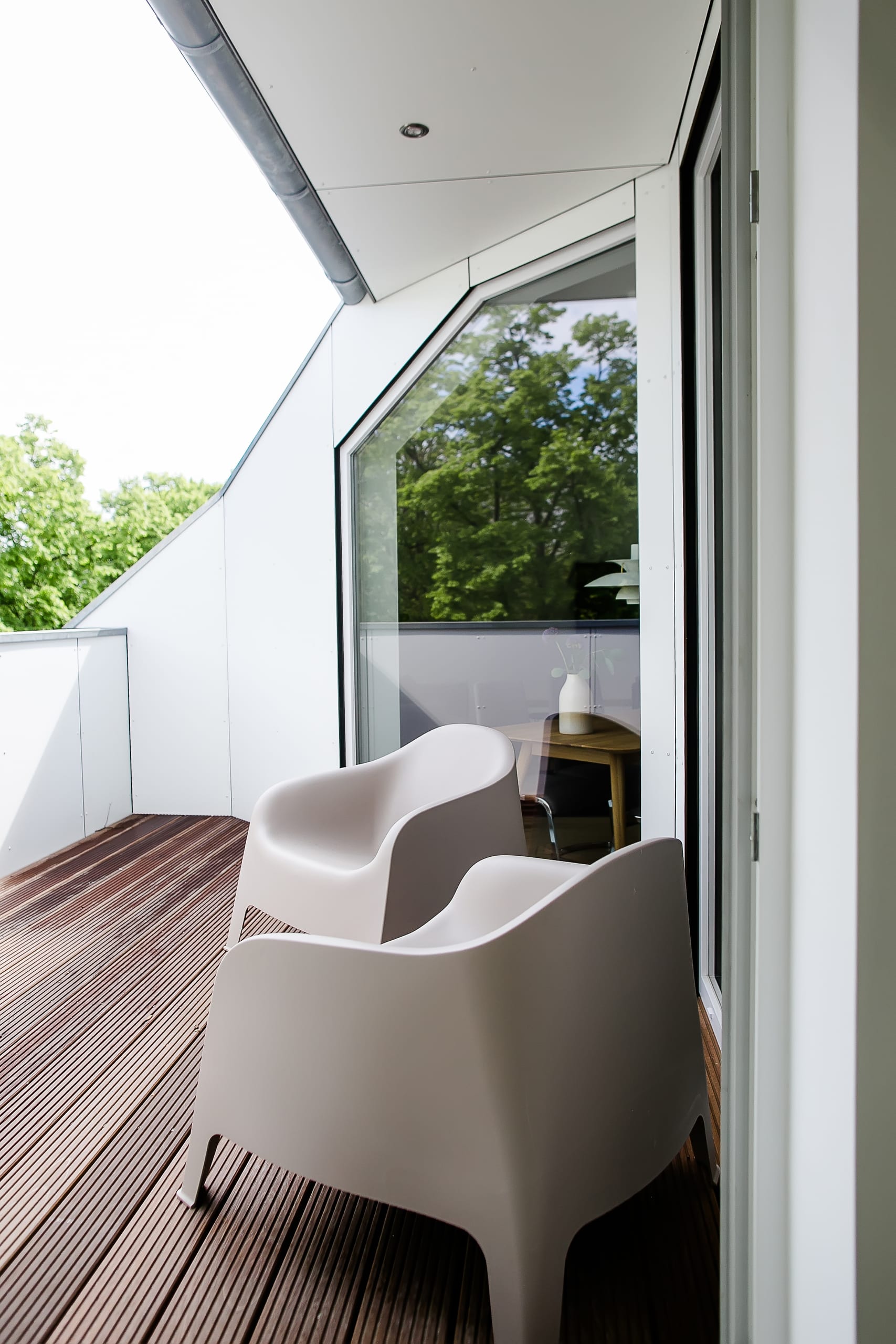 Balkon mit Holzplankenboden, zwei beigen Stühlen und Fensterfront zu grünen Bäumen