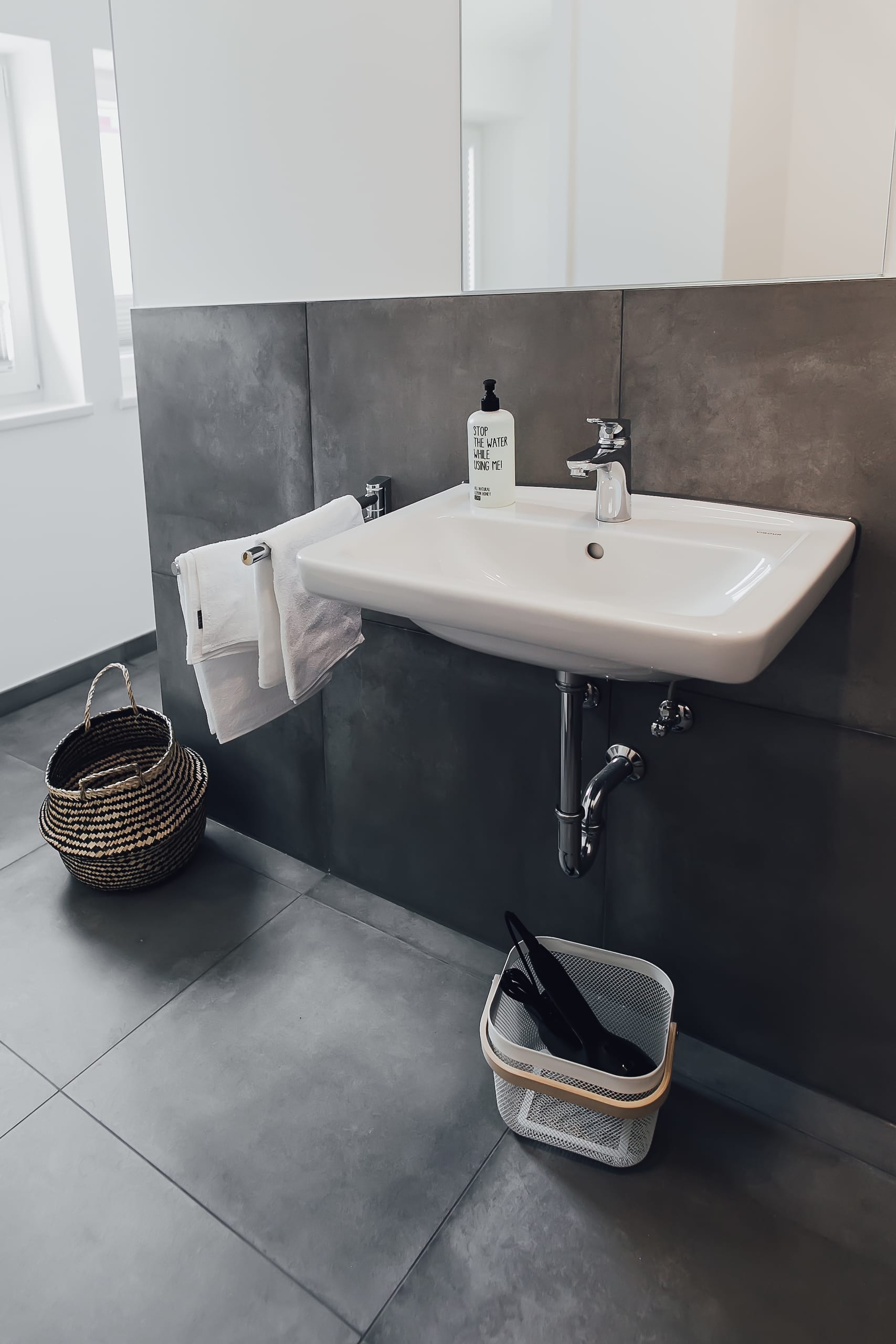 Badezimmer mit weißem Waschbecken, dunklen Fliesen, links ein geflochtener Korb