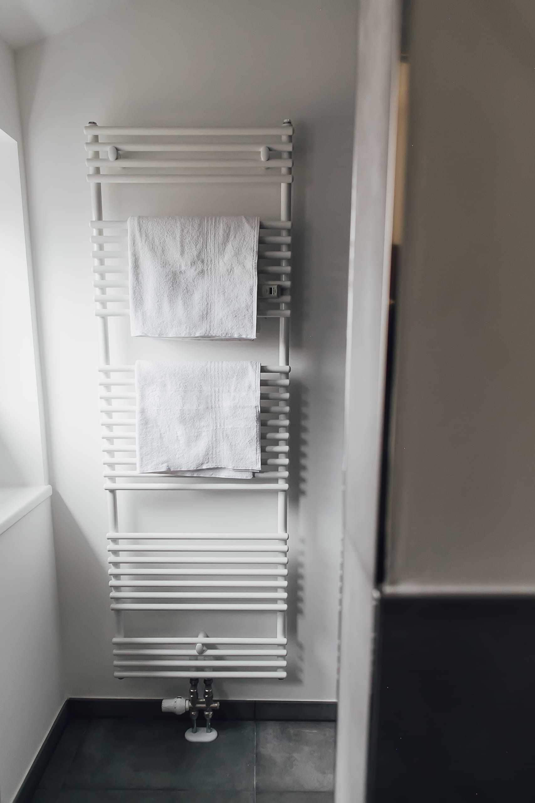 Weißer Handtuchheizkörper, zwei gefaltete Handtücher, an heller Wand auf dunklem Fliesenboden