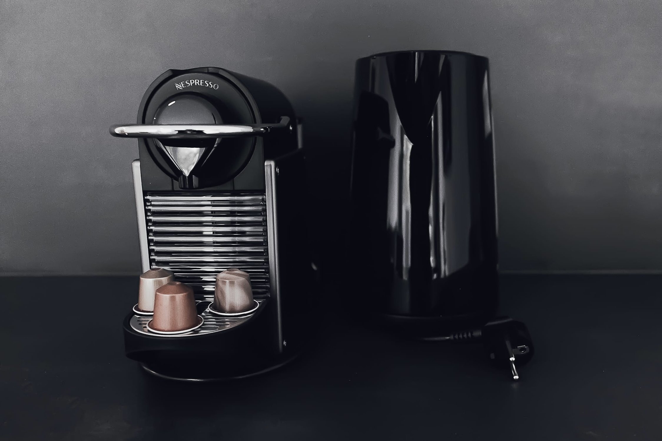 Schwarze Nespresso-Maschine mit Kapselhalterung, drei Kapseln und schwarzer Thermoskanne daneben