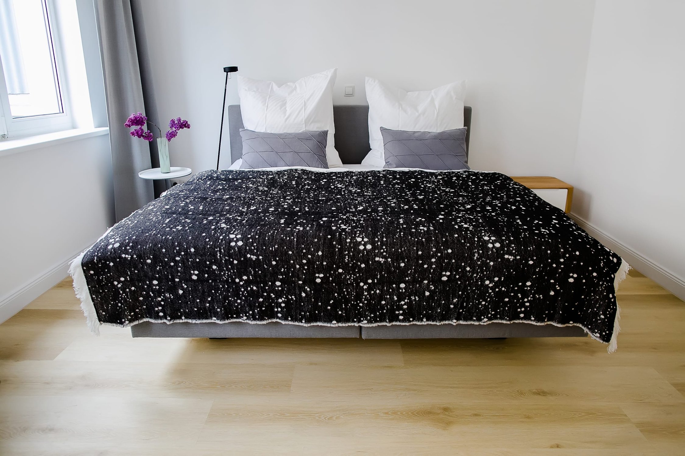 Schlafzimmer mit Doppelbett, schwarzer Tagesdecke, Holzfußboden und Fenster mit grauem Vorhang
