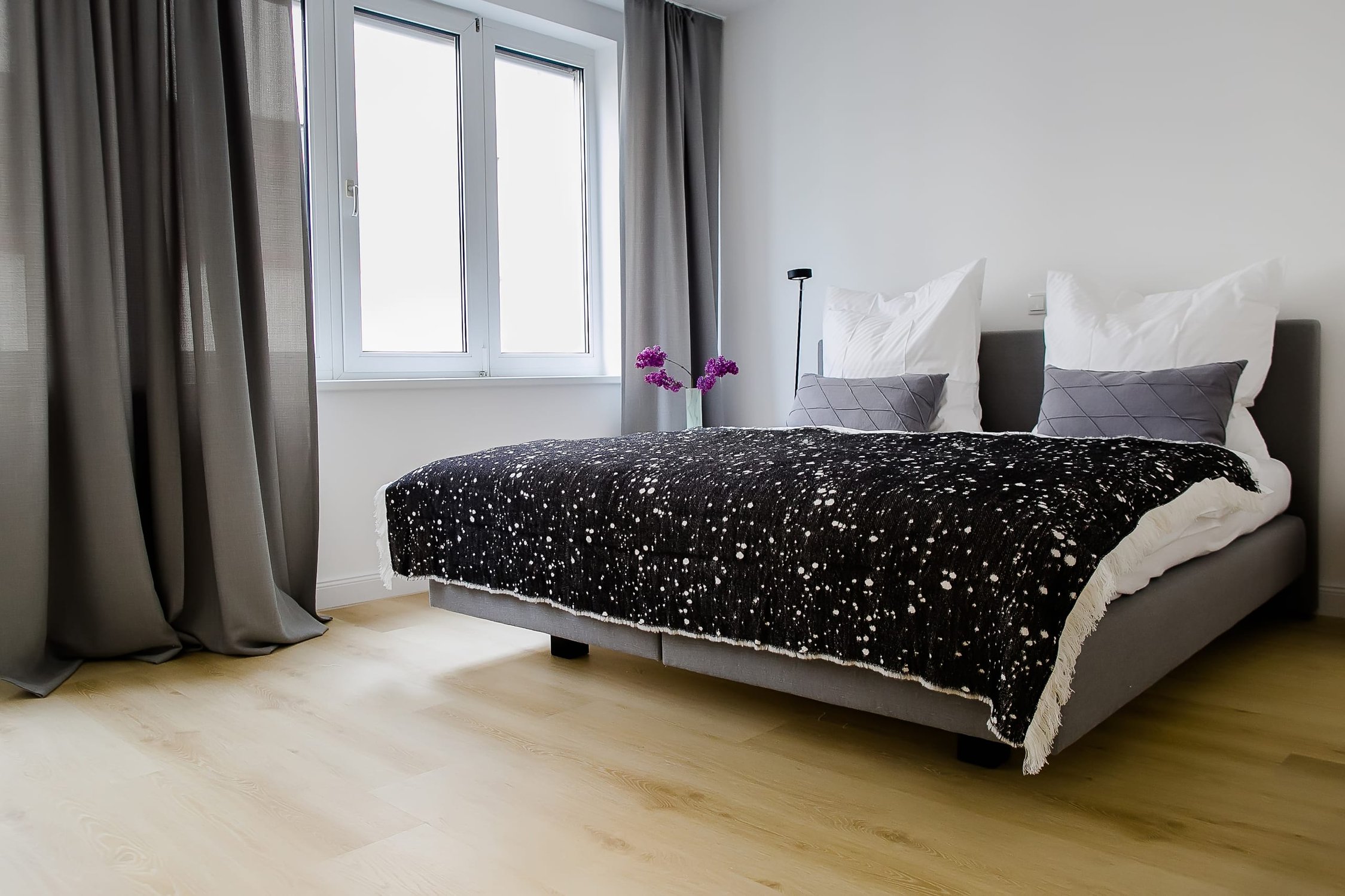 Schlafzimmer, großes graues Bett mit gemusterter schwarzer Decke, großes Fenster, Holzfußboden