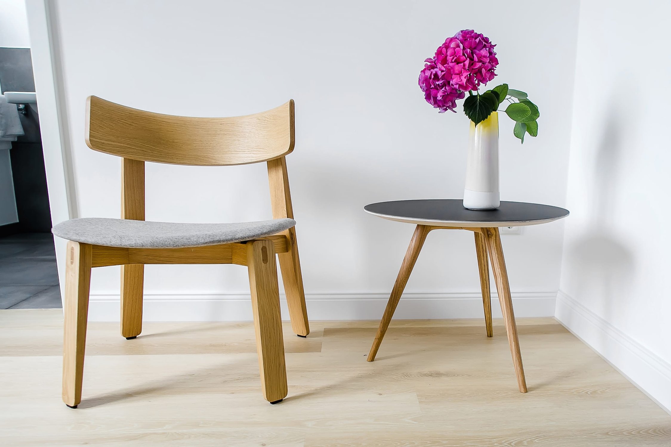 Holzstuhl mit grauem Polster neben Beistelltisch mit pinken Blumen in weißer Vase