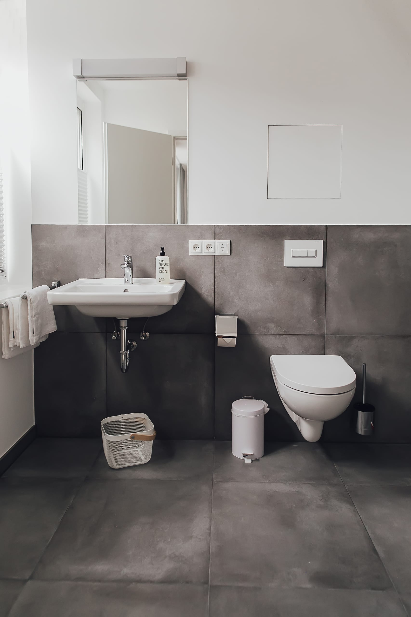 Badezimmer mit grauen Fliesen, weißem Waschbecken und Toilette, Spiegel über dem Waschbecken