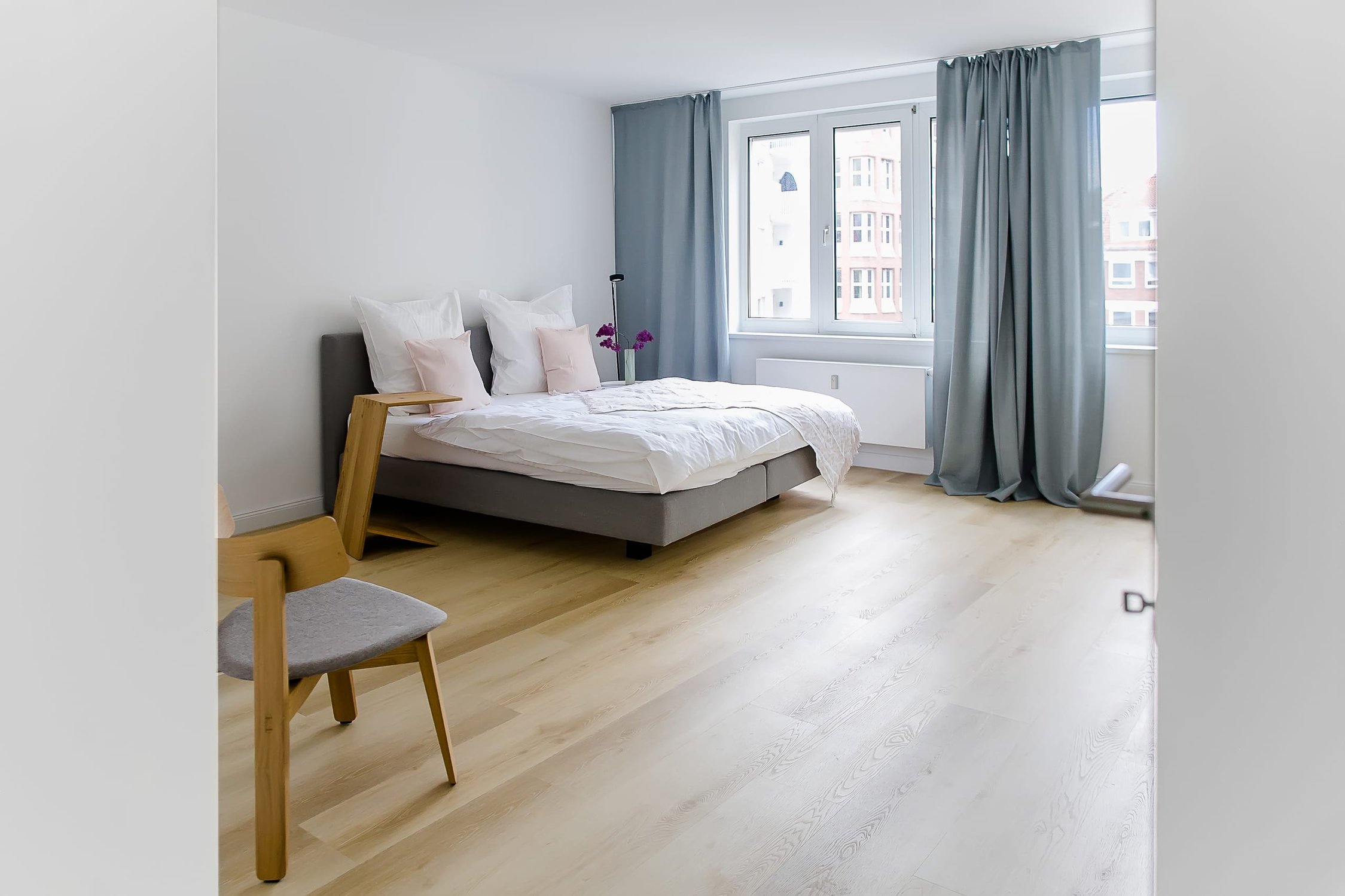 Schlafzimmer mit weißem Bett und blauen Vorhängen, Holzboden, einem Stuhl und Fenster