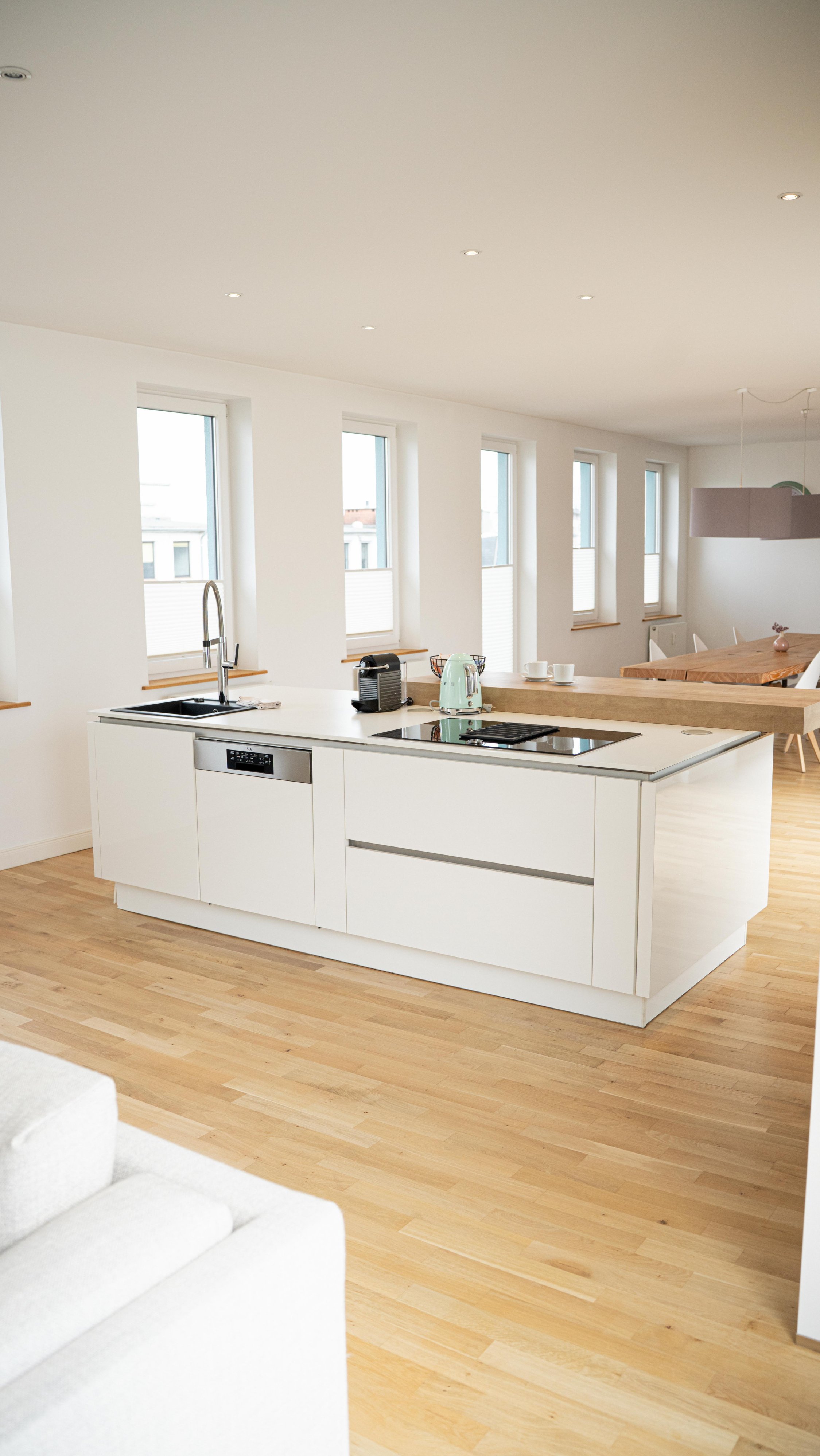 Moderne offene Küche mit weißer Inselküche, Edelstahlgeräten, großen Fenstern mit Holzrahmen, hellem Holzboden und Essbereich im Hintergrund