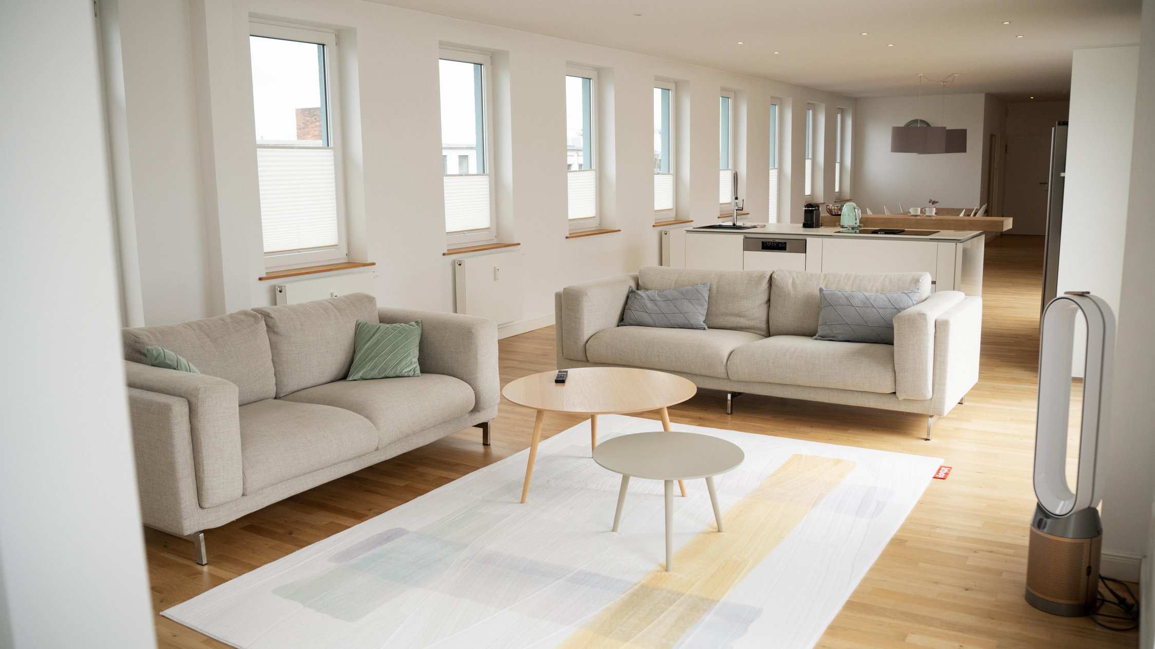 Offenes Wohnzimmer mit zwei beigen Sofas, runden Holzcouchtischen, großen Fenstern mit Jalousien, hellem Holzboden und offener Küche im Hintergrund