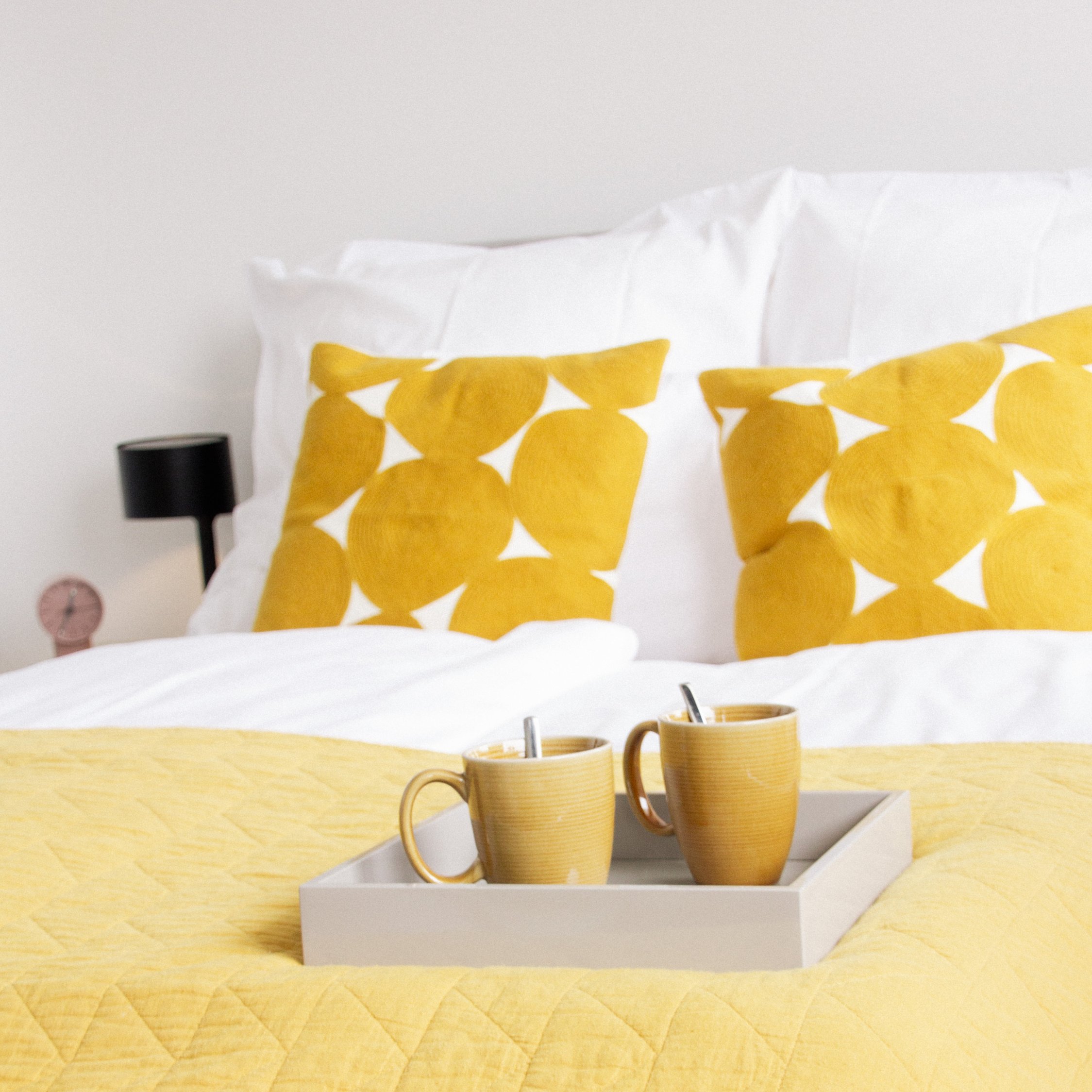 Nahaufnahme eines Bettes mit gelben gemusterten Kissen mit geometrischen Dreiecken, weißer Bettwäsche und zwei goldenen Tassen auf einem Tablett