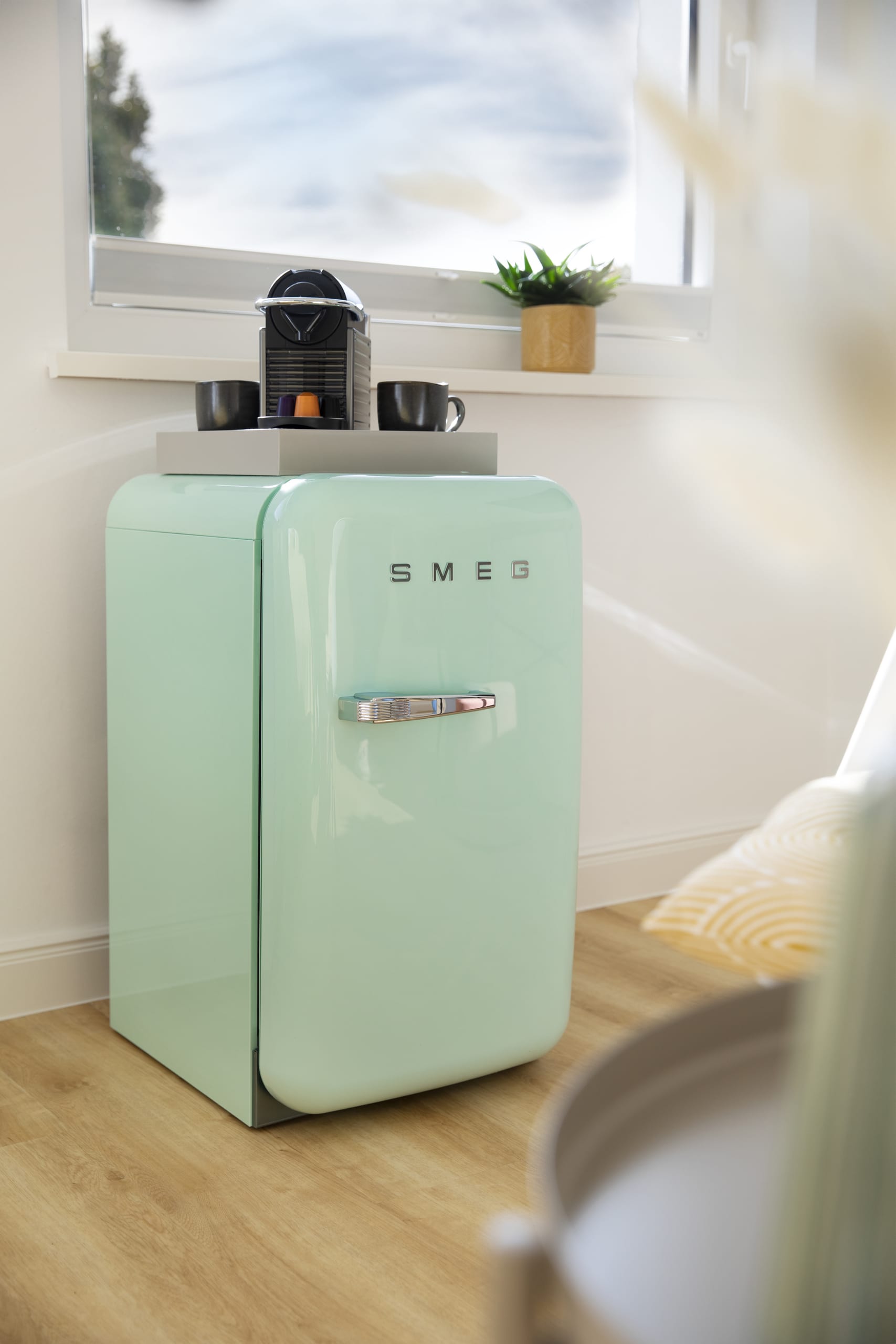 Minikühlschrank in Mintgrün mit integrierter Kaffeemaschine und Tassen unter Fenster, heller Holzfußboden