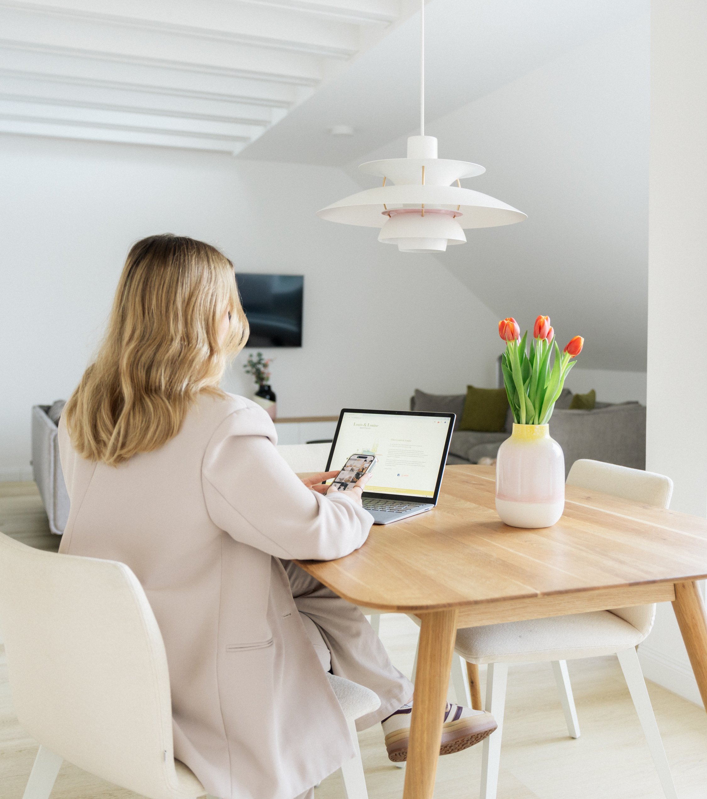 Frau in beigem Blazer arbeitet an einem hellen Holztisch mit Laptop und Smartphone unter weißer Pendelleuchte, rote Tulpen in weißer Vase auf dem Tisch